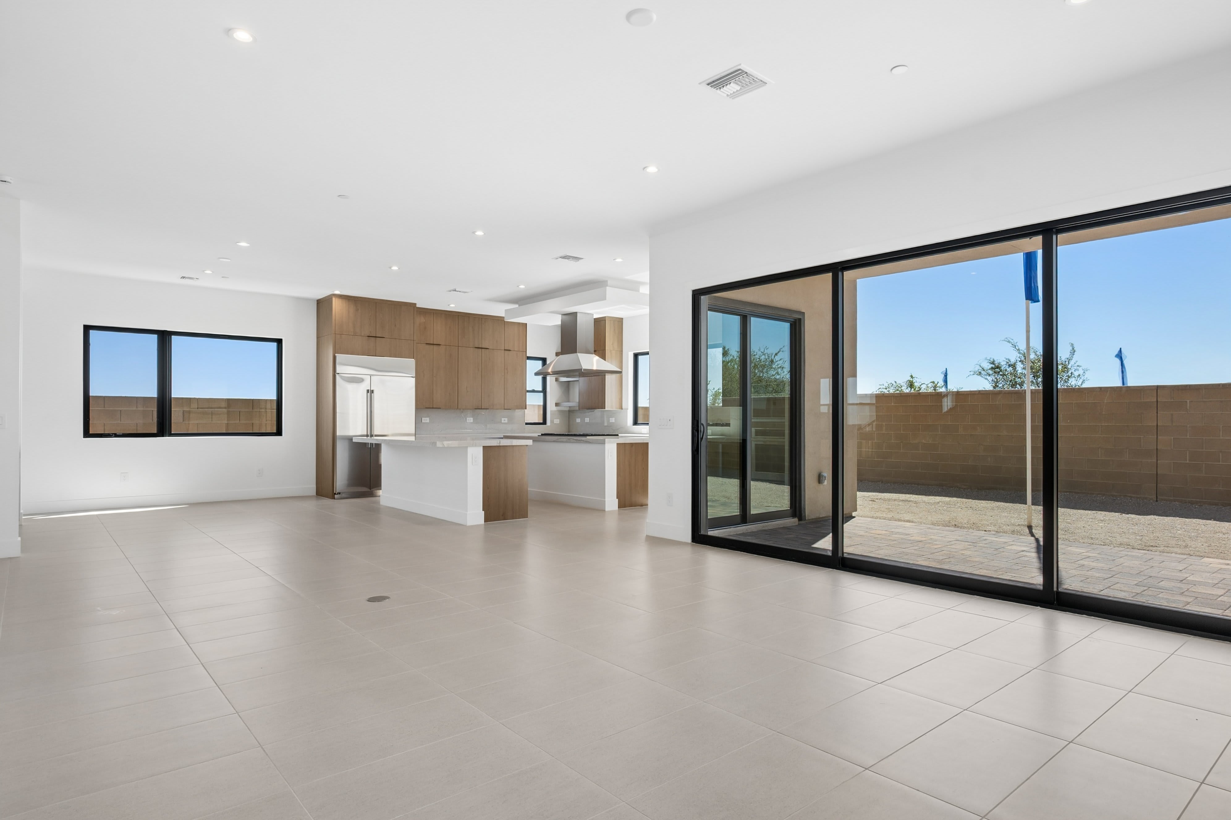 A spacious, empty room with light-colored tile flooring leads to a modern kitchen and large sliding glass doors that open to a fenced backyard.