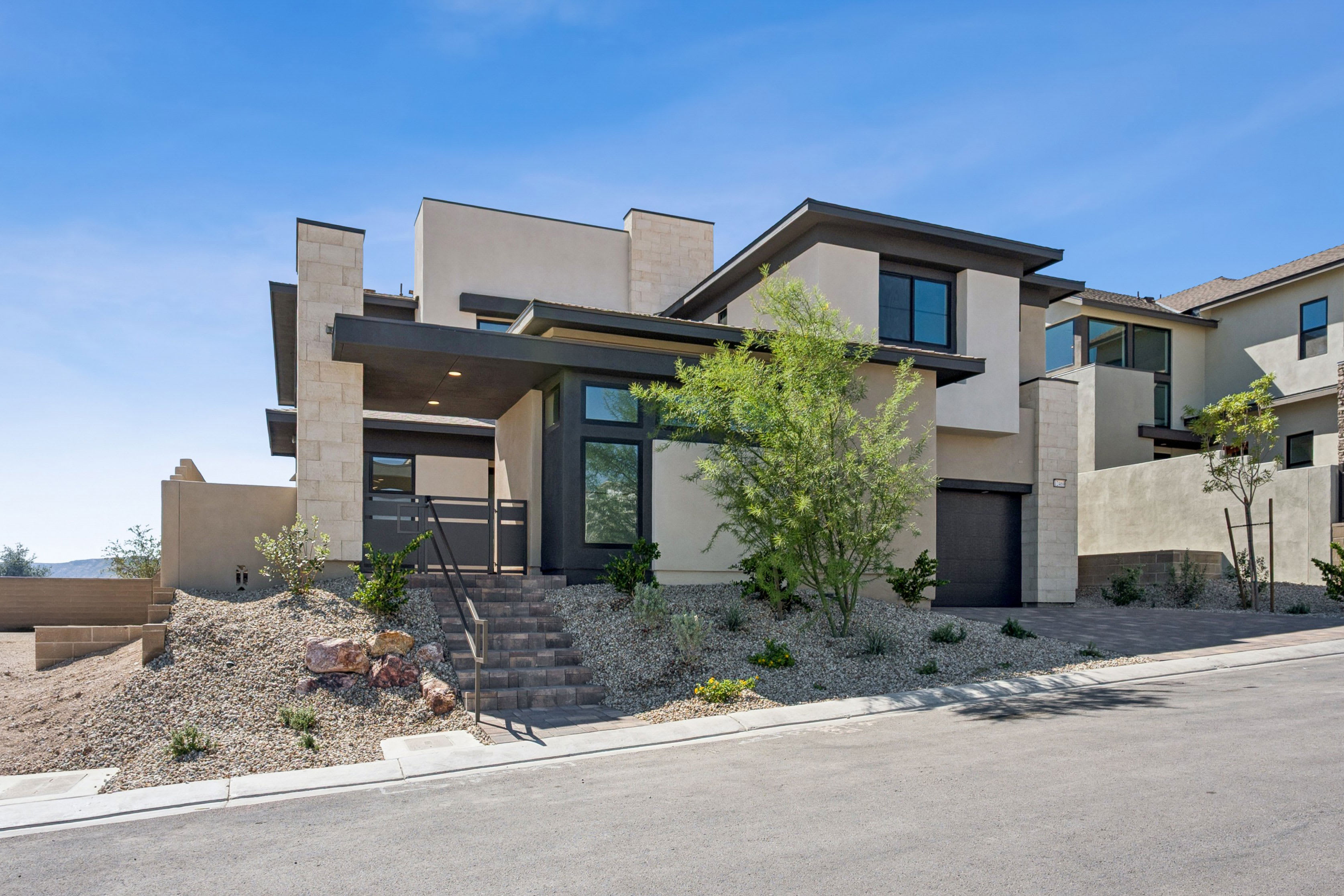 A modern, multi-level beige house with dark trim stands behind a landscaped yard of gravel and greenery under a clear blue sky.