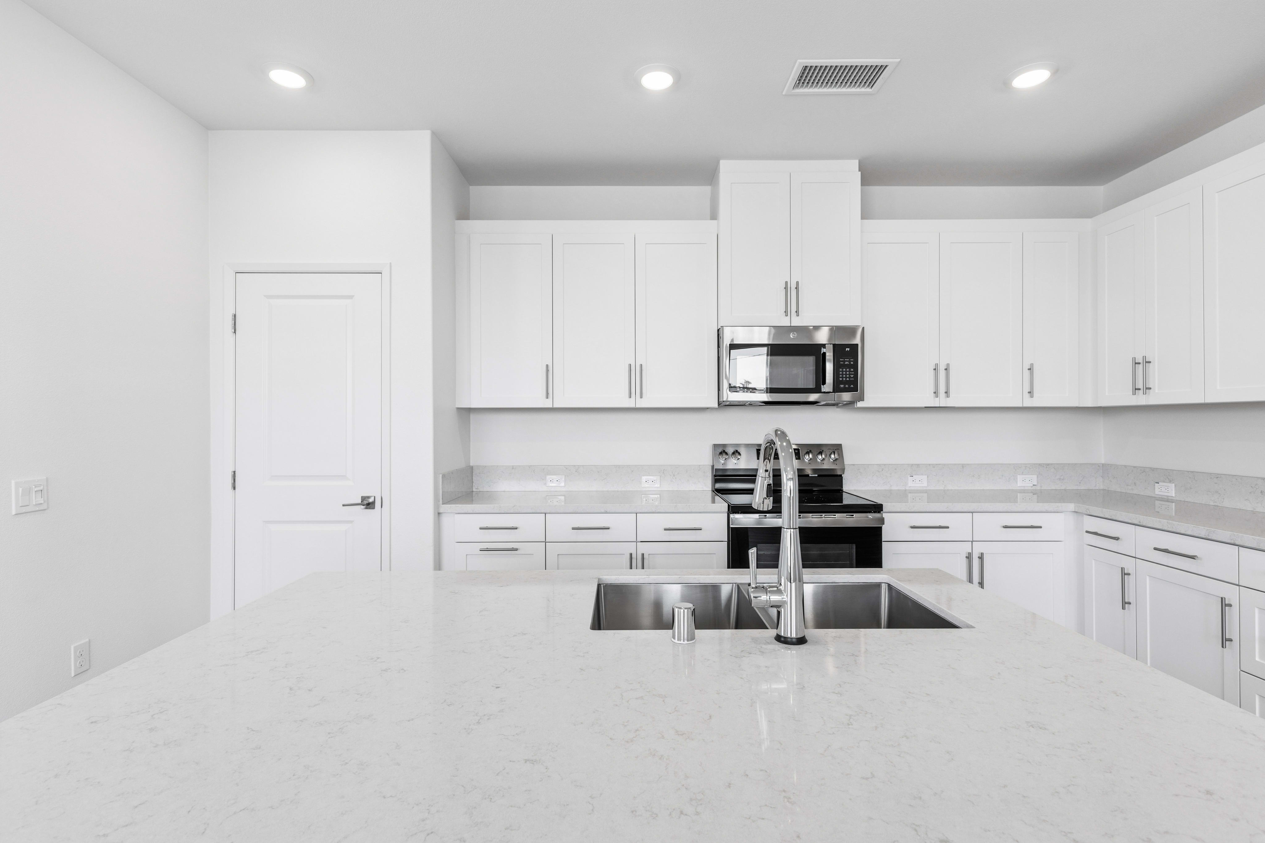 A white quartz kitchen island with a stainless steel sink sits in the foreground of a modern white kitchen featuring matching cabinetry and appliances.