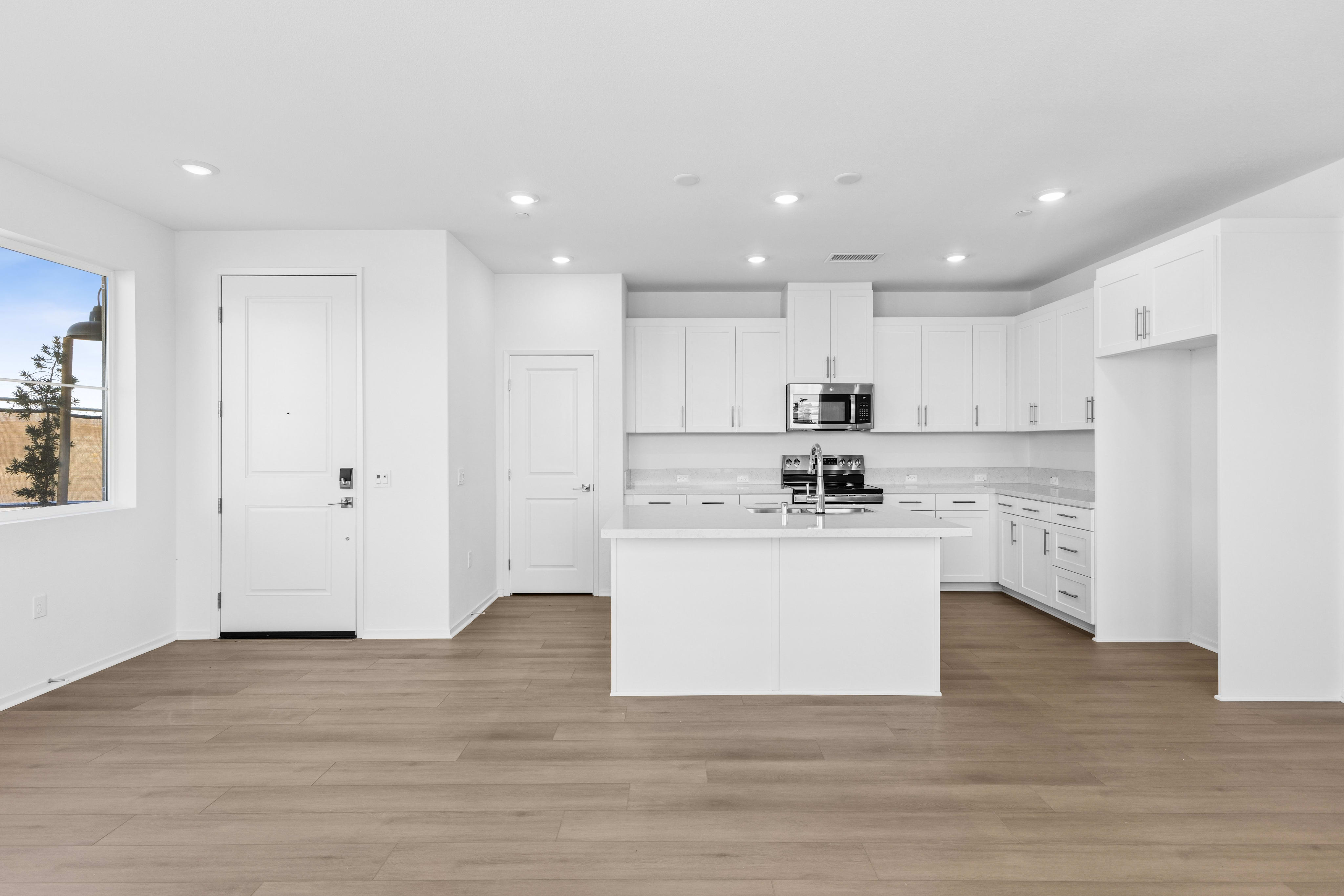 An empty room with light wood flooring leads to a modern white kitchen with a central island and white cabinetry in the background.