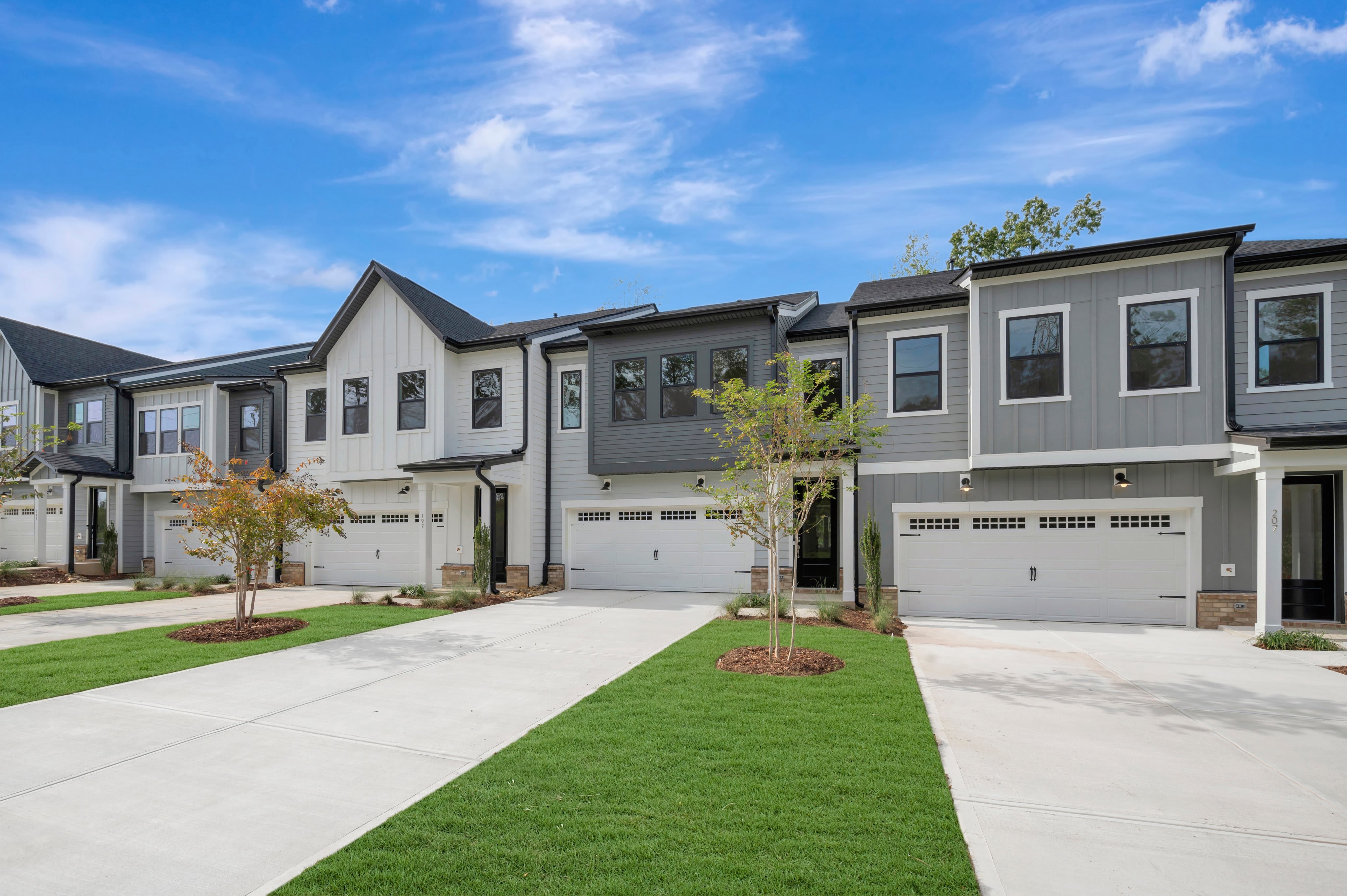 Modern townhomes with white driveways and green lawns sit under a bright blue sky with wispy clouds.