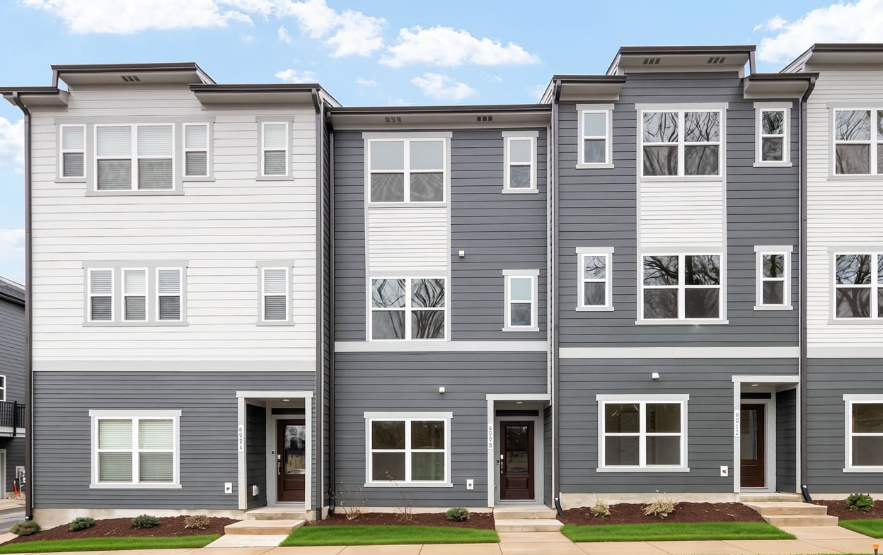 Modern grey and white townhouses stand under a bright blue sky with scattered clouds, fronted by small patches of green grass and mulch.
