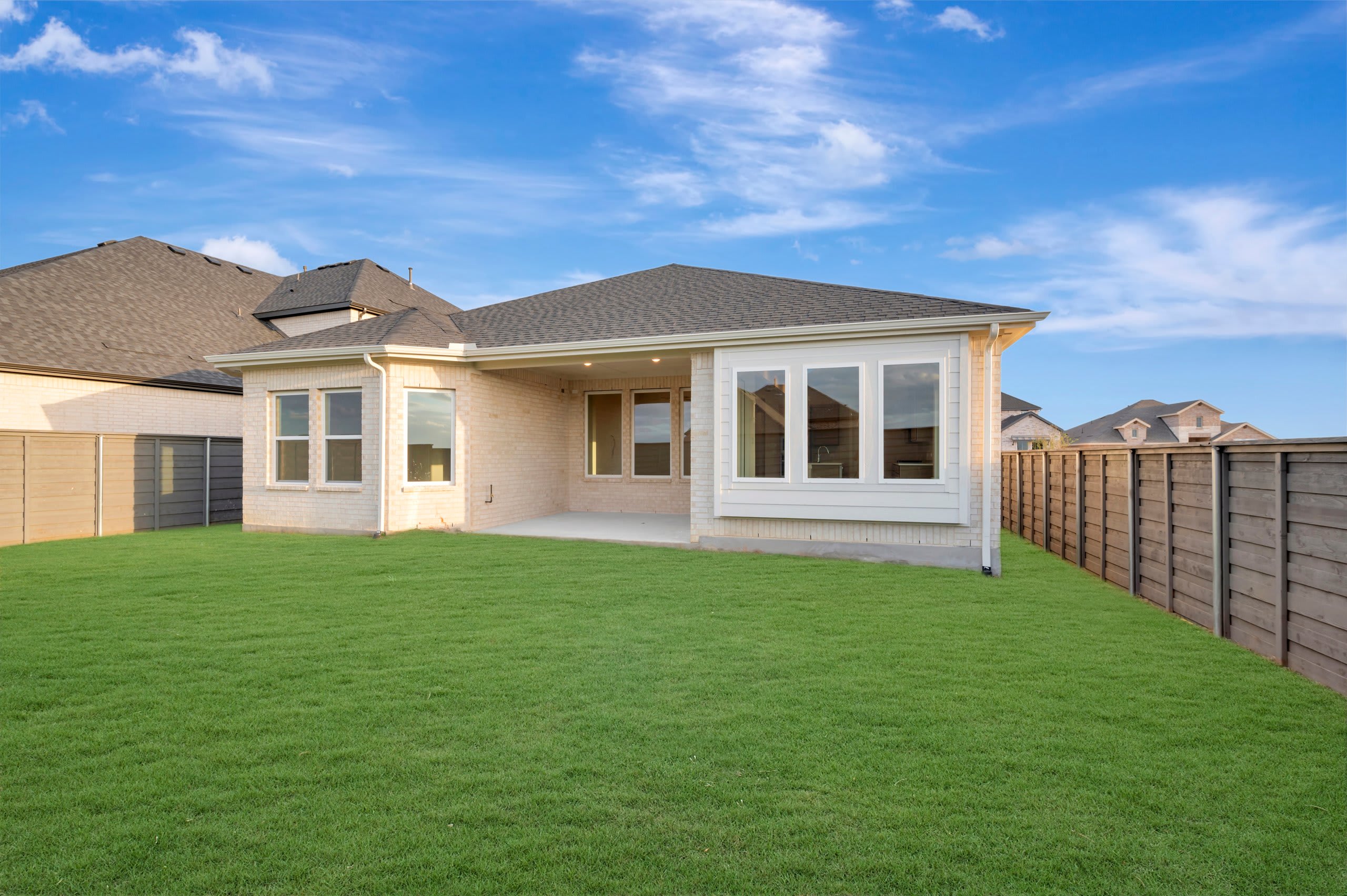A lush green lawn fills the foreground, leading to a modern light-colored brick house set against a bright blue sky with wispy clouds.