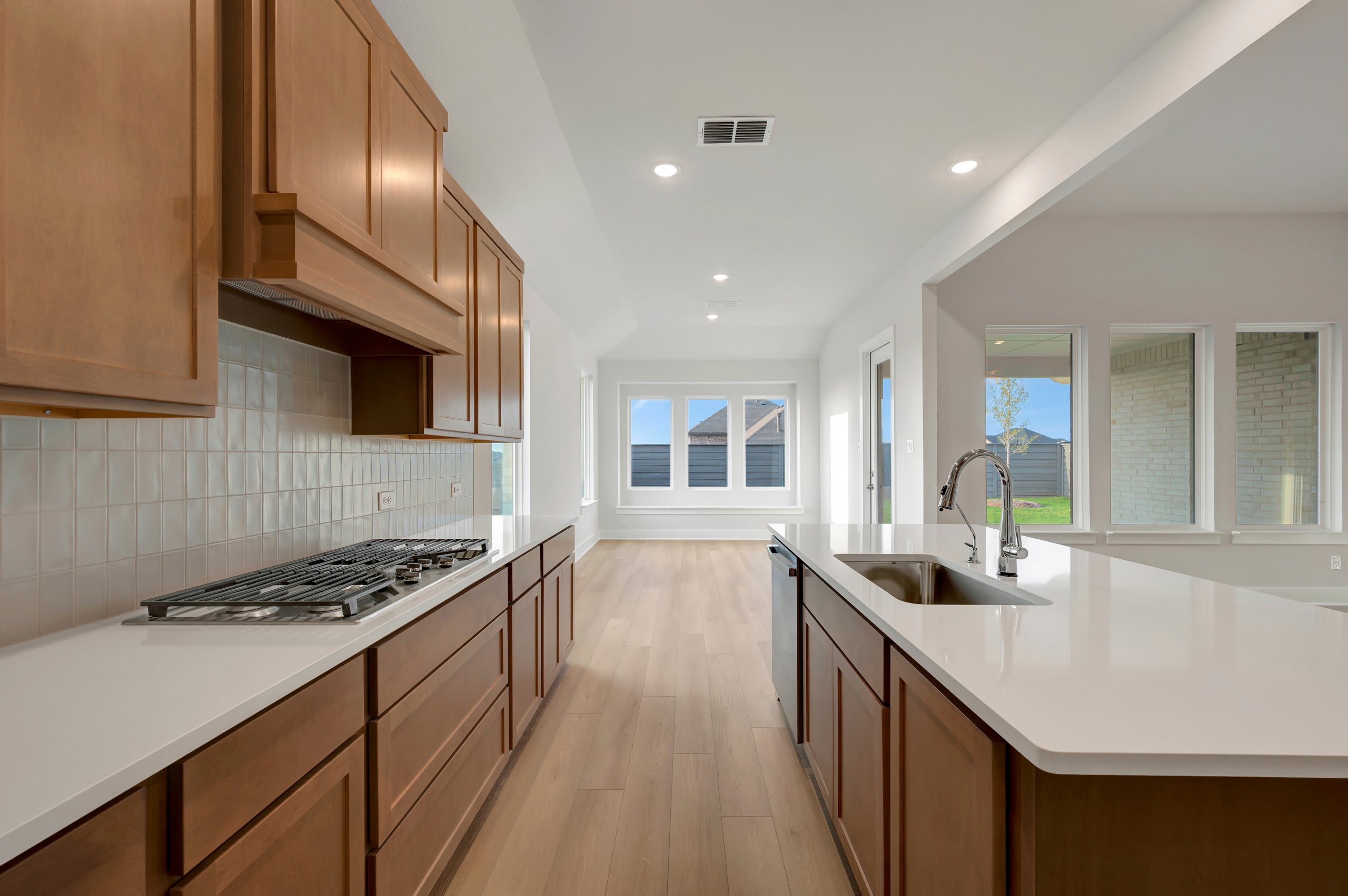 A modern kitchen features white countertops and wooden cabinetry in the foreground, leading toward a bright living area with large windows in the background.
