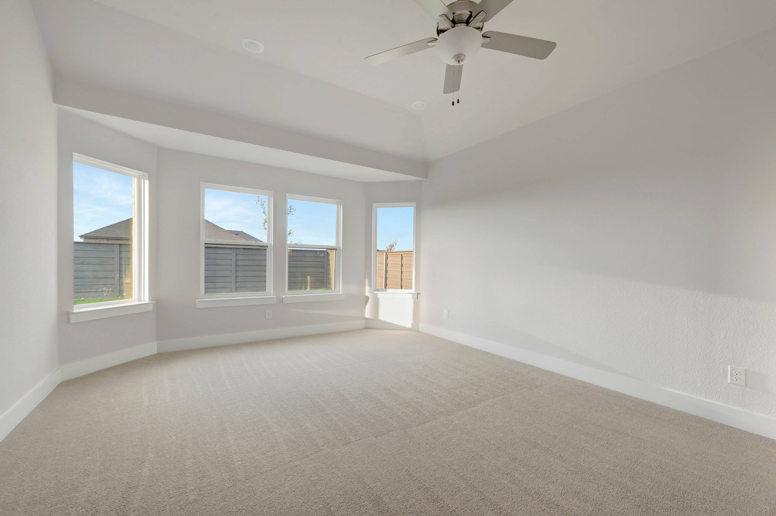 An empty room with beige carpeting in the foreground and white walls with several windows and a ceiling fan in the background.