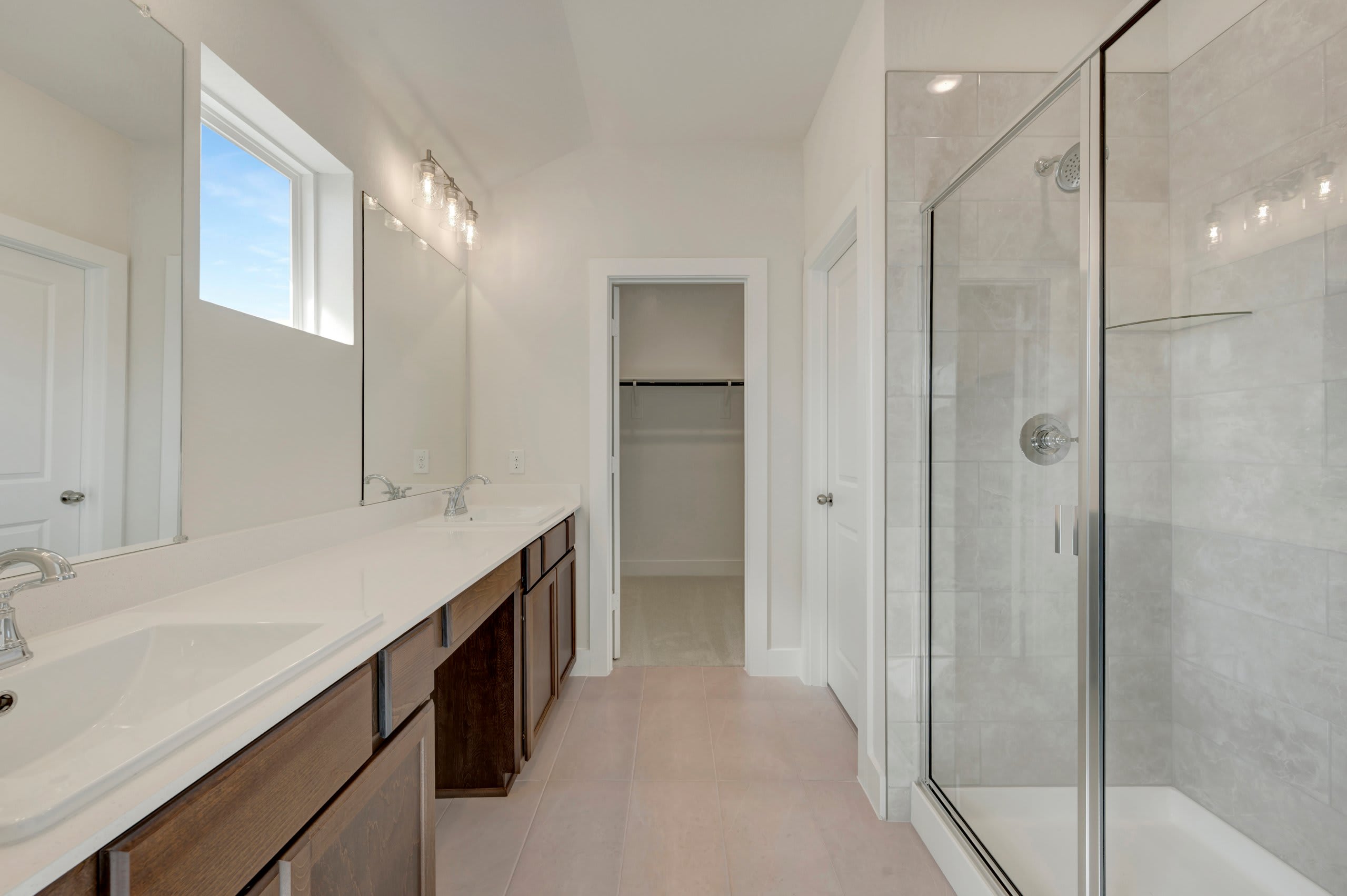 A modern bathroom features a long white vanity with dark wood cabinets in the foreground and a glass-enclosed shower and open closet in the background.