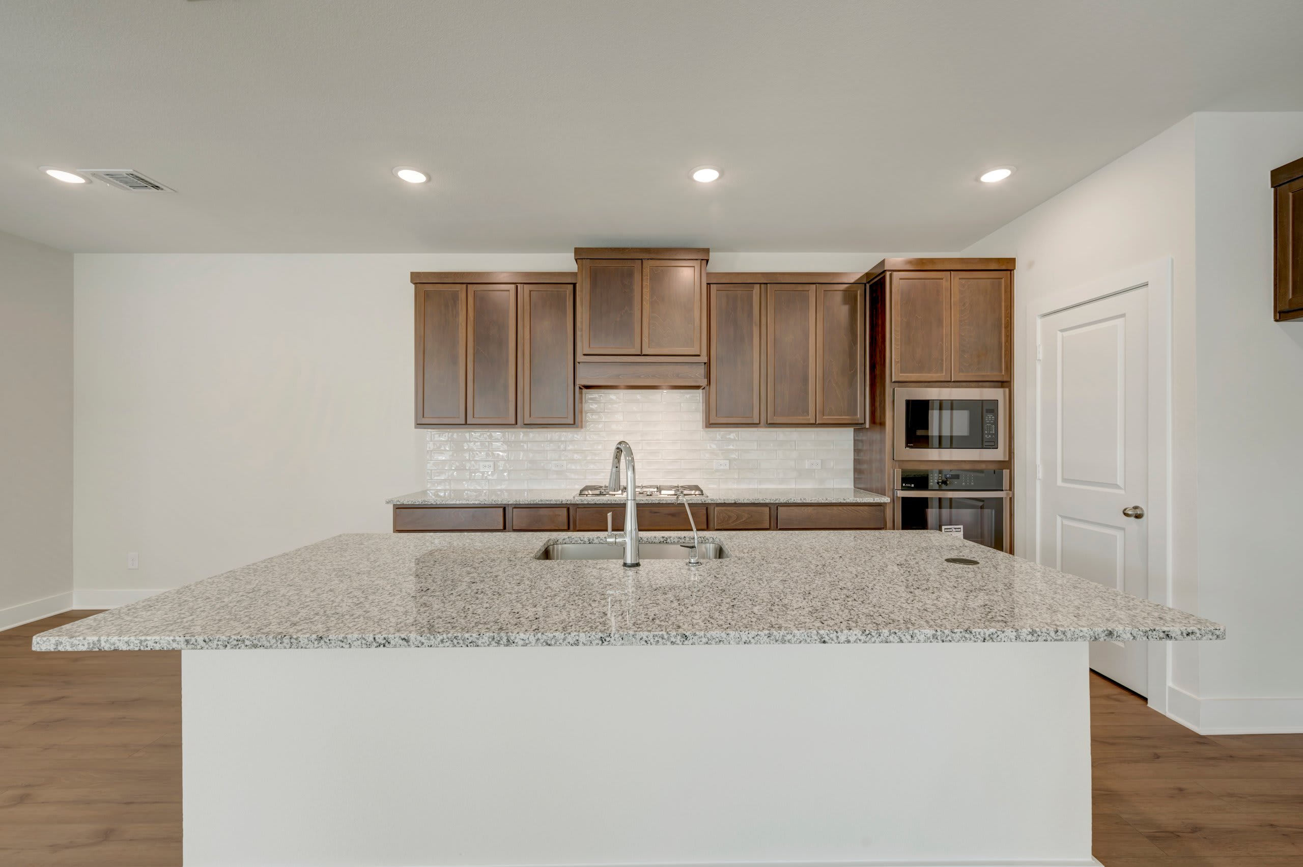 A granite kitchen island with a sink sits in the foreground of a room featuring brown wooden cabinets and white walls in the background.