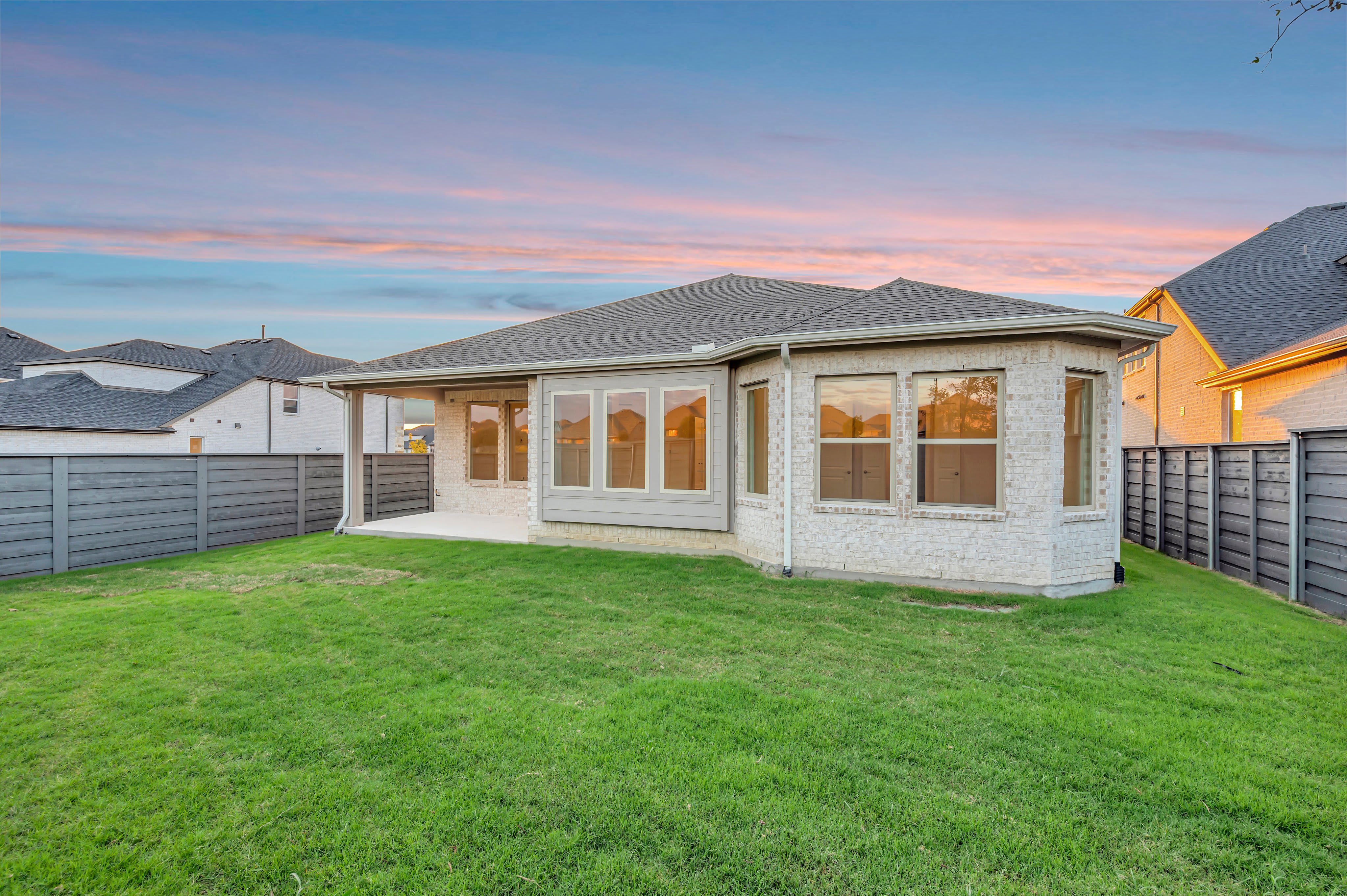 A lush green lawn fills the foreground, leading to a modern white brick house set against a colorful sunset sky.