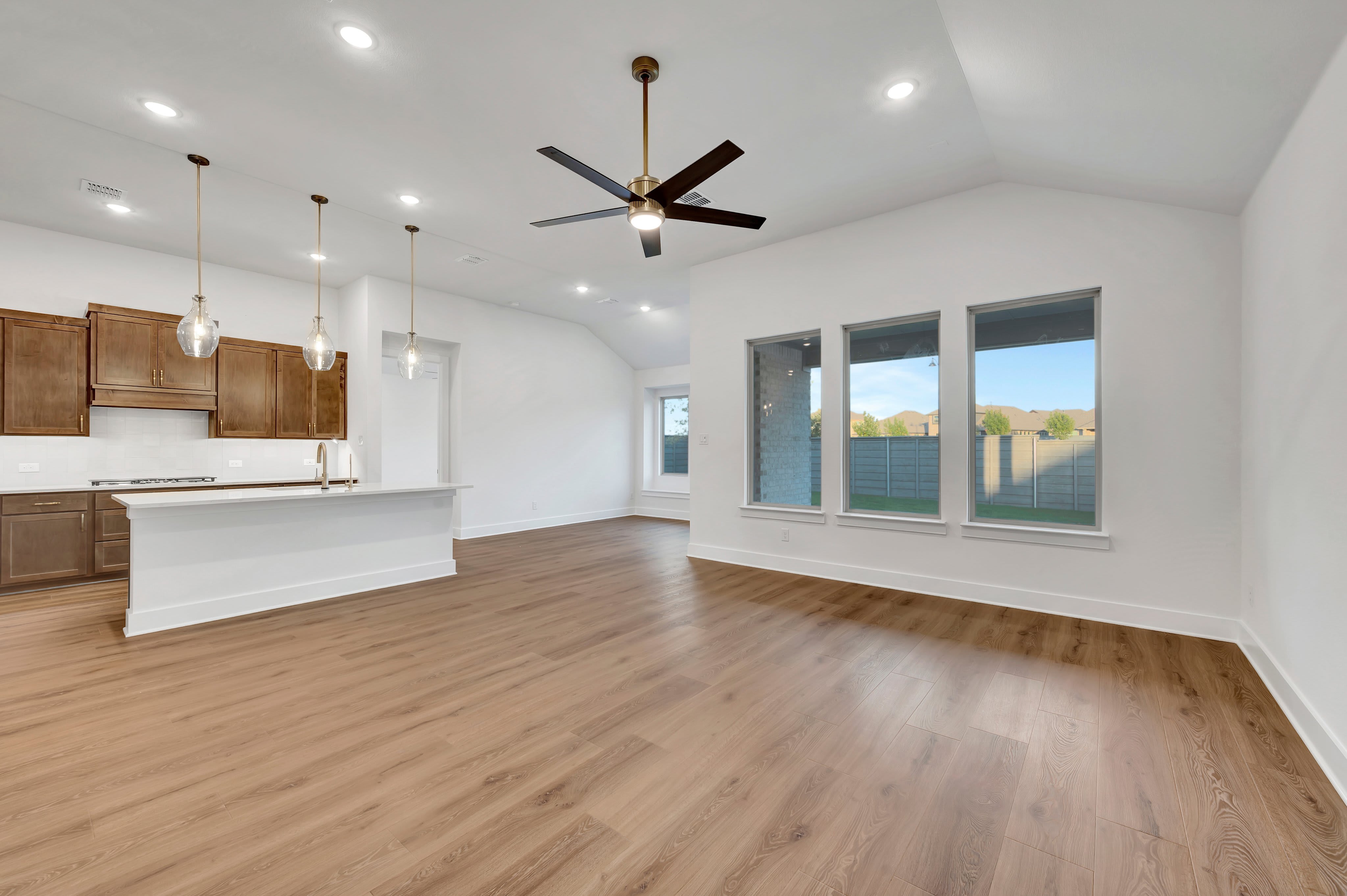 An open-concept living area with light wood flooring and a white kitchen island in the foreground leads to a bright room with a ceiling fan and large windows overlooking a backyard in the background.
