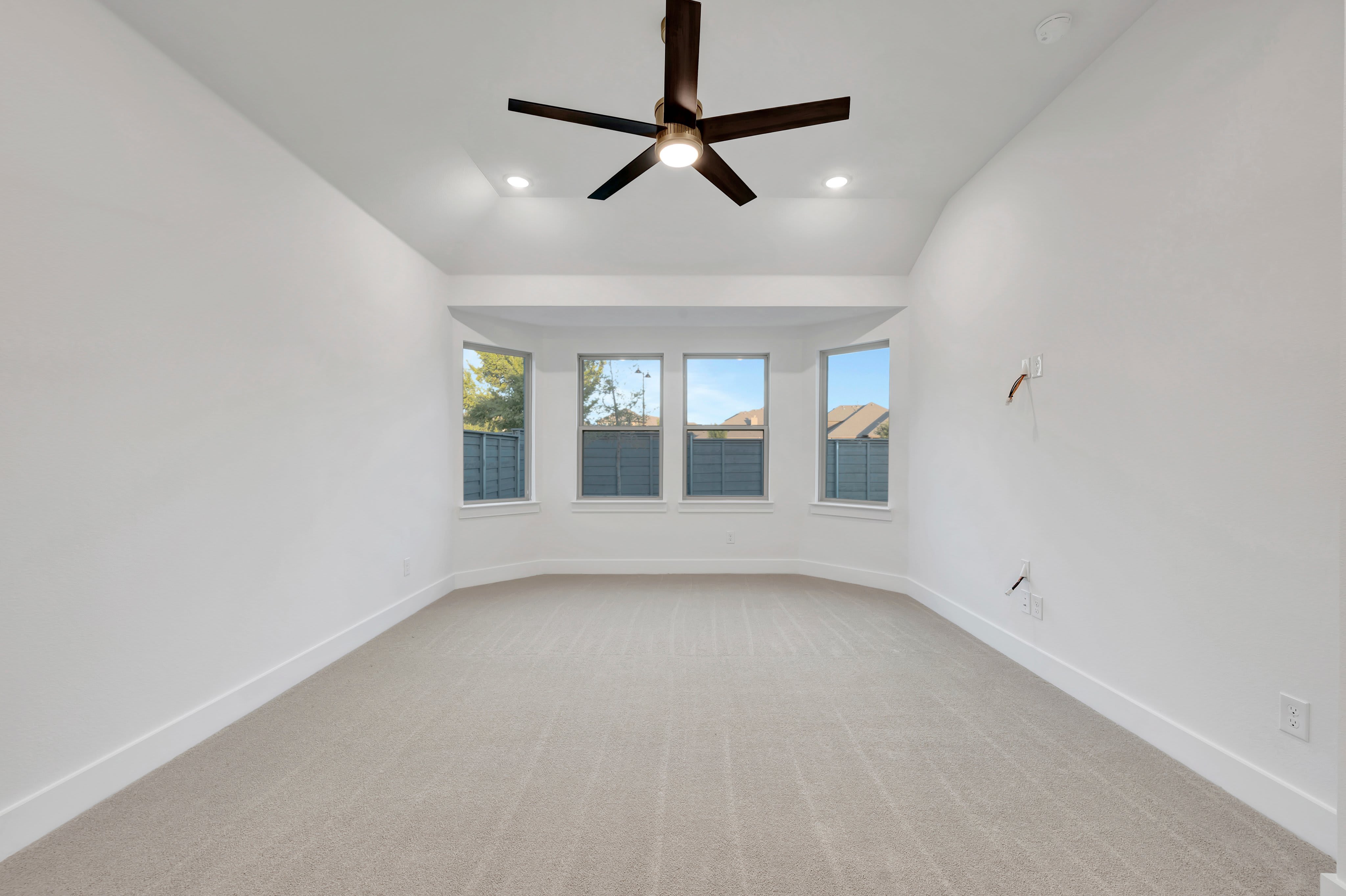 An empty room with beige carpeting and white walls features a dark ceiling fan in the foreground and a bay window overlooking a neighborhood in the background.