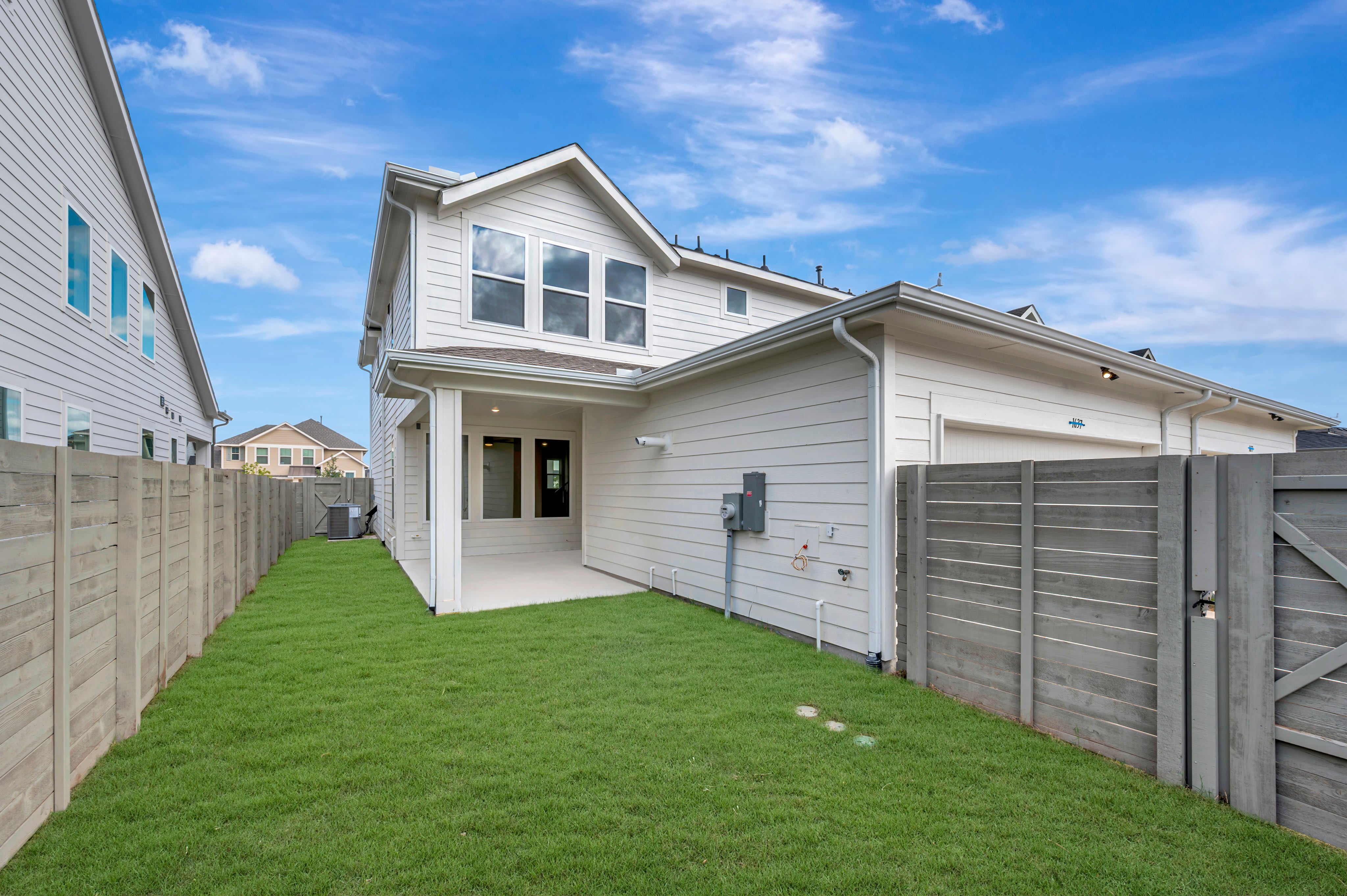 A green grassy backyard is enclosed by wooden fences, leading to a white two-story house under a bright blue sky with wispy clouds.