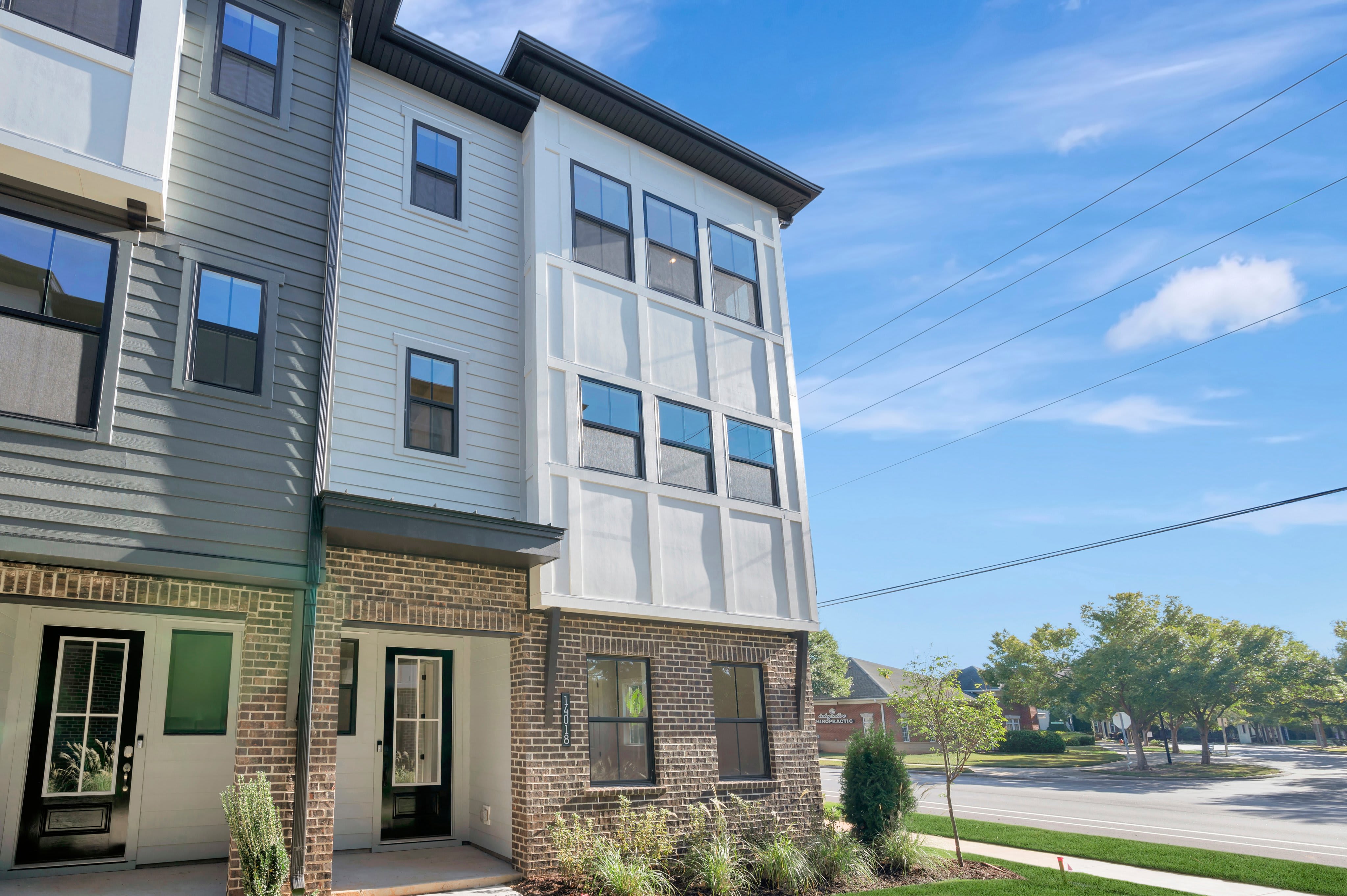 A modern multi-story residential building with grey and white siding and brick accents stands against a clear blue sky with distant green trees and a road.