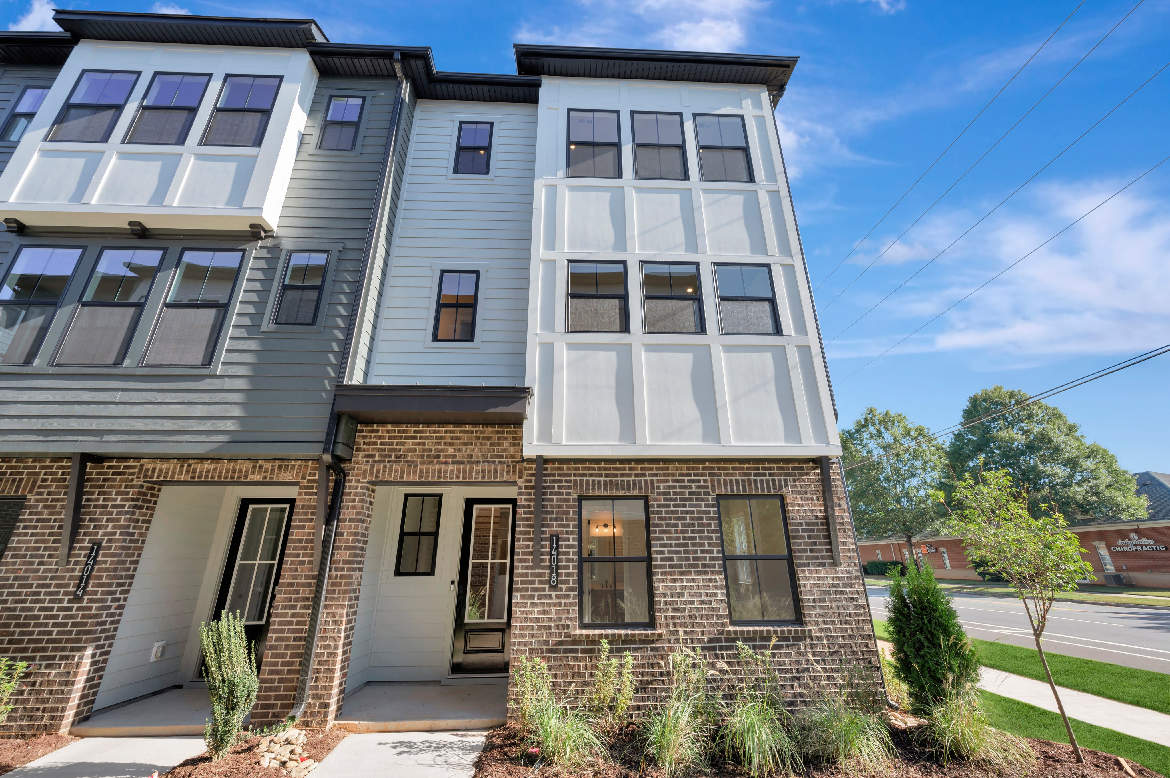 A modern multi-story townhouse with a mix of brick, grey, and white siding stands in the foreground against a bright blue sky with light clouds.