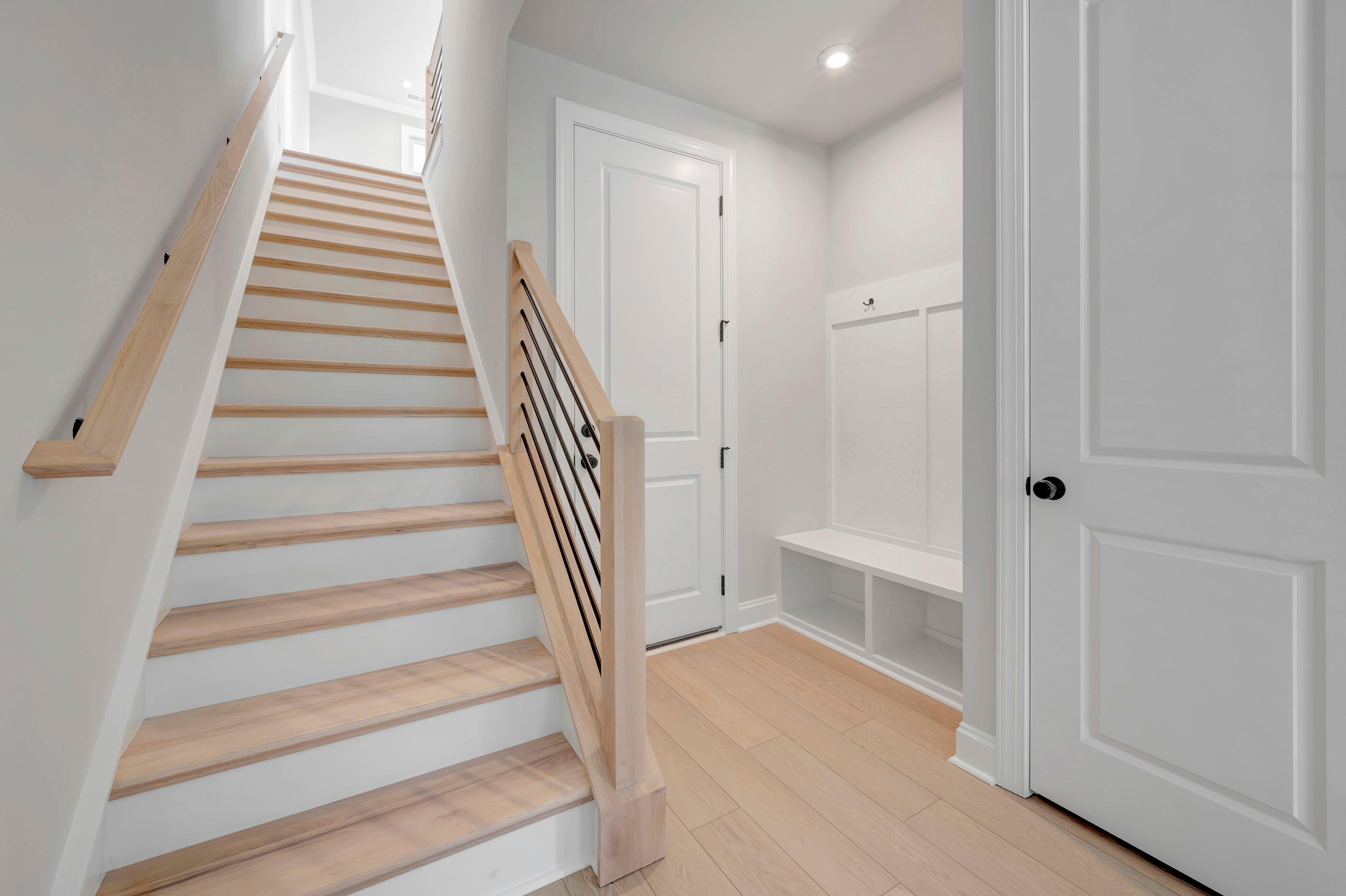A light-colored wooden staircase with white risers leads upward in the foreground, while a white hallway with a built-in bench and closed doors fills the background.
