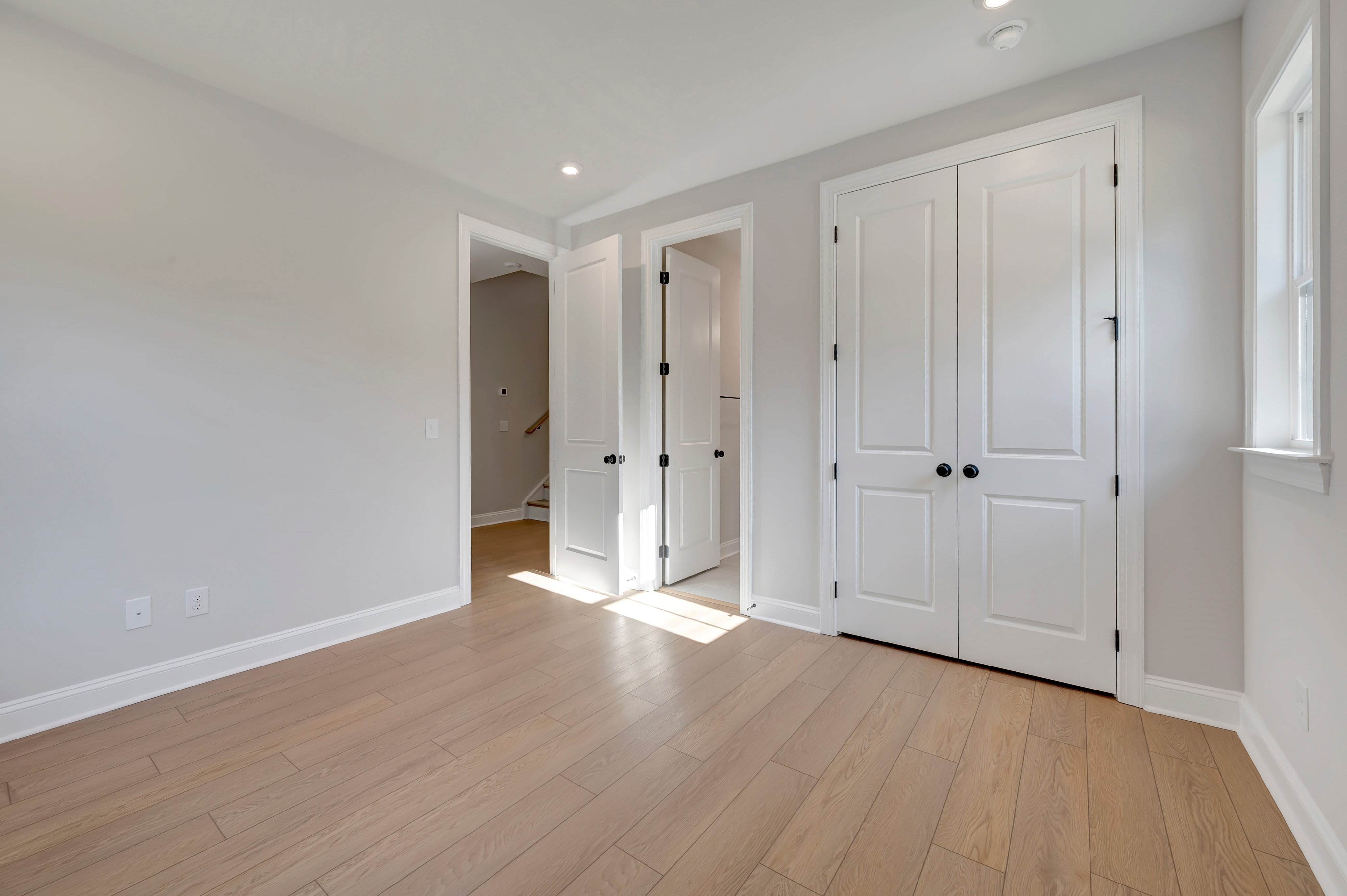 Light-colored hardwood floors fill the foreground of an empty room with white walls and several white doors leading to other spaces in the background.