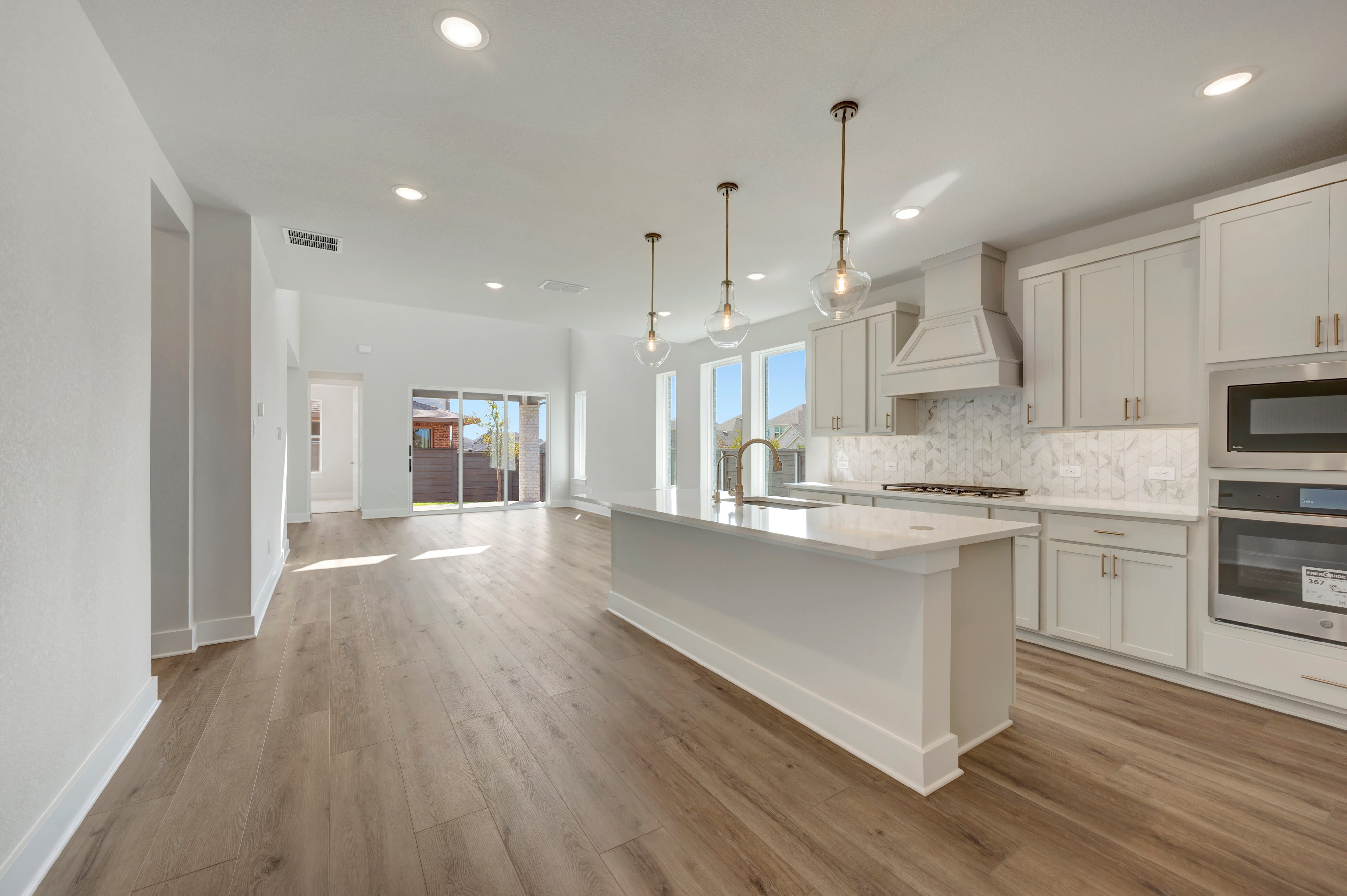 A modern white kitchen with a large center island and hardwood floors opens into a bright, spacious living area with sliding glass doors in the background.