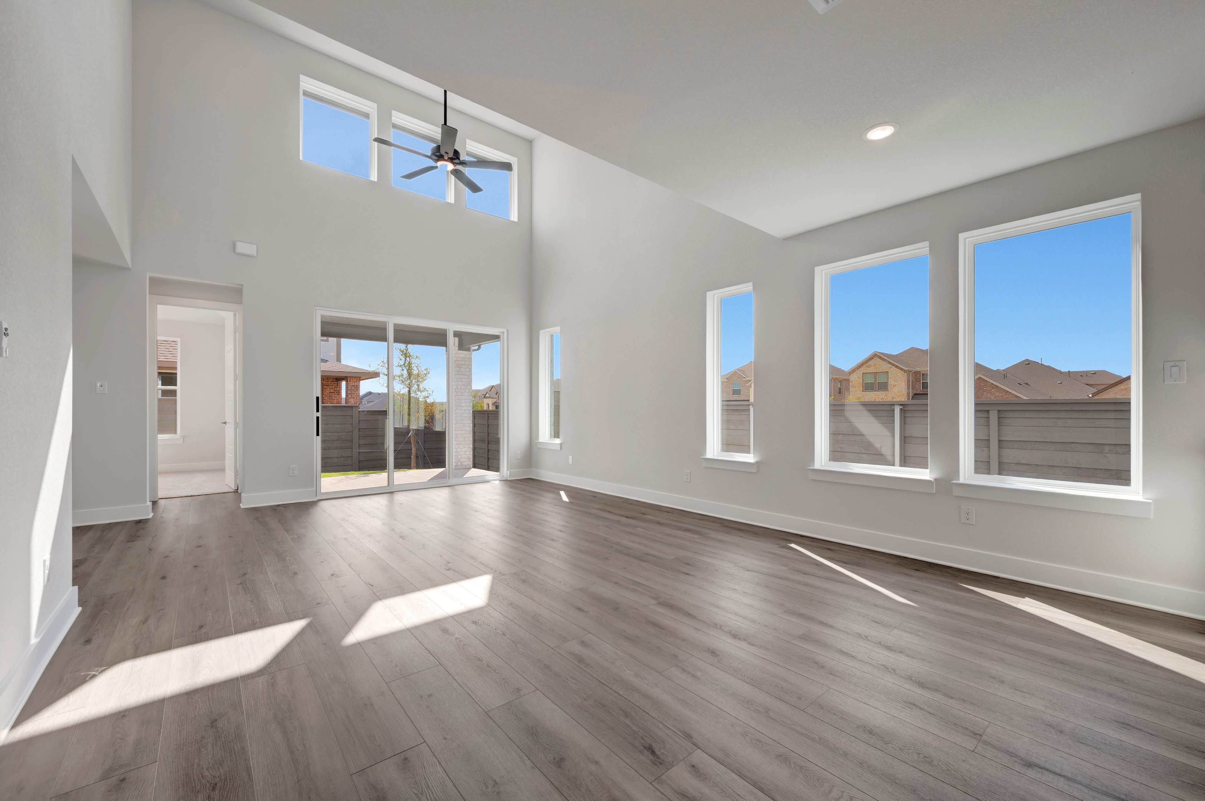 An empty room with grey wood flooring in the foreground and large windows overlooking a residential neighborhood in the background.