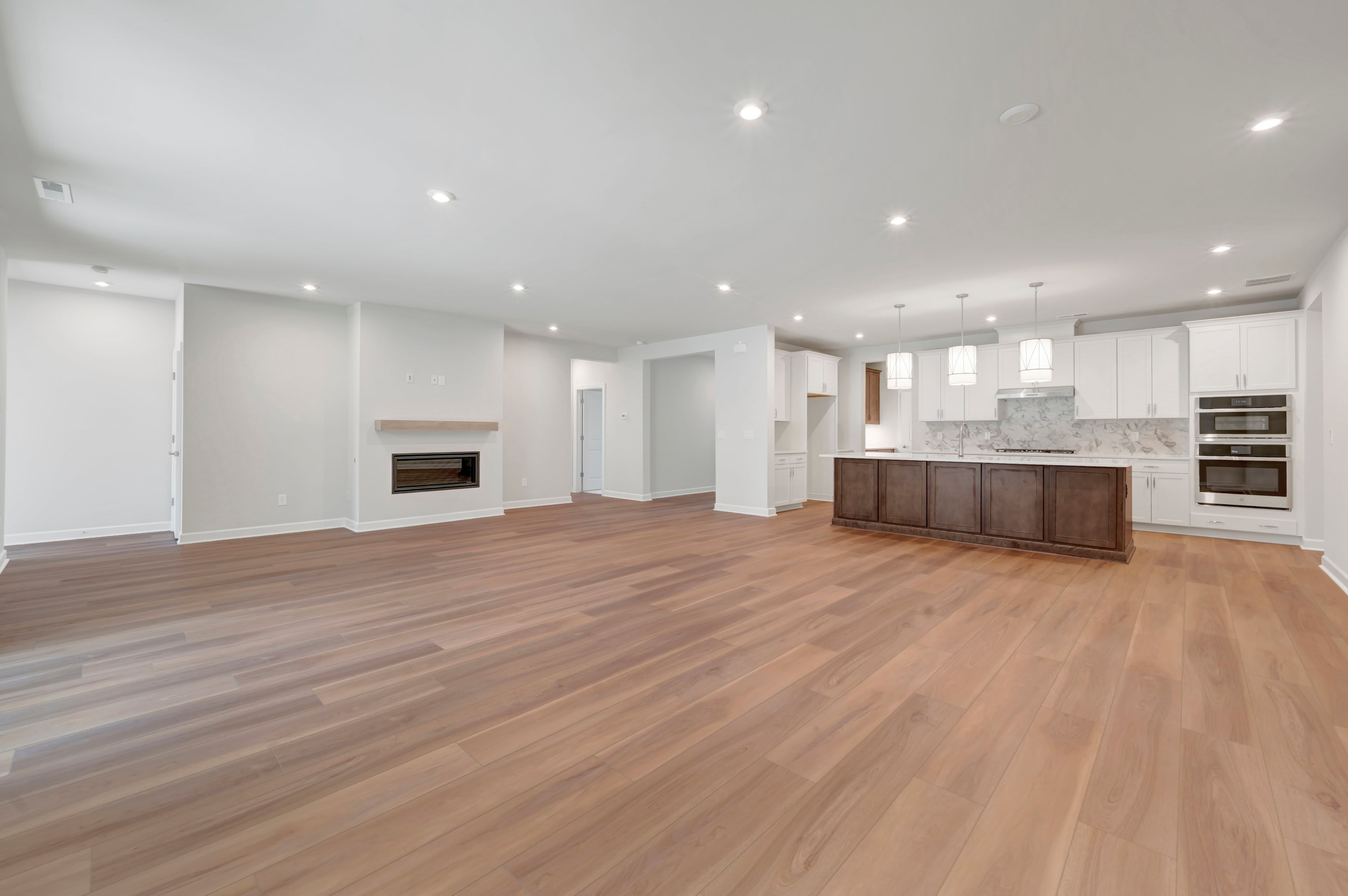 An open-concept living area with light hardwood floors in the foreground leads to a modern kitchen and a fireplace in the background.