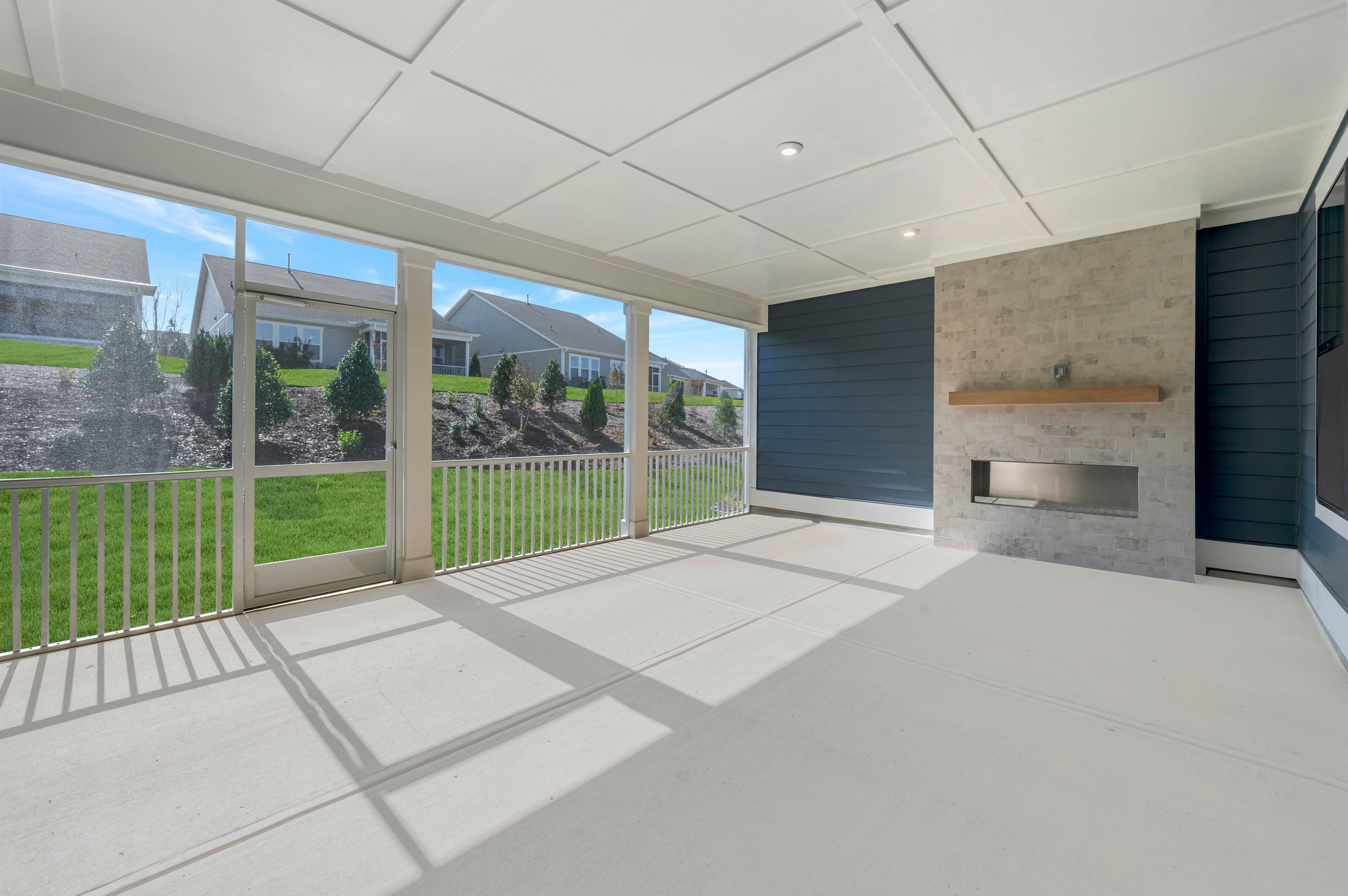An empty screened-in porch with a modern fireplace and white ceiling overlooks a green lawn and neighboring houses.