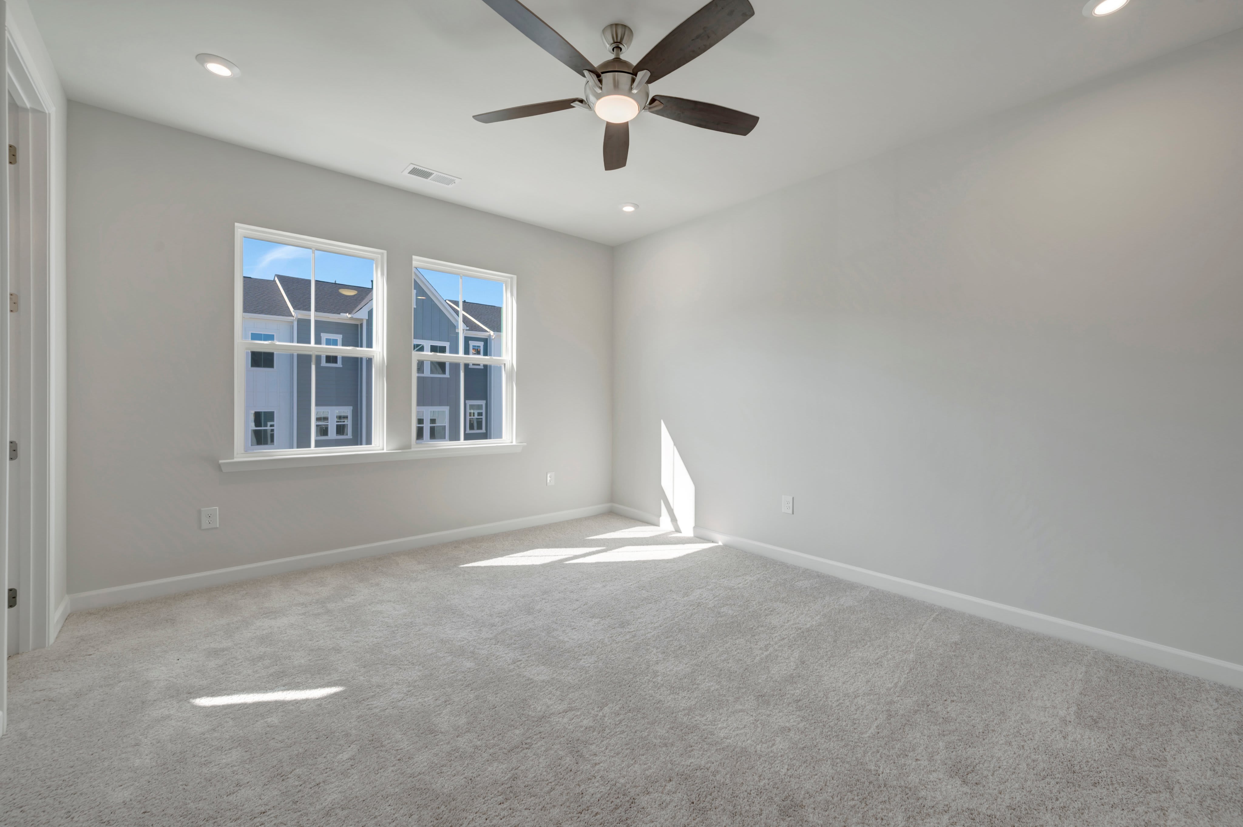 An empty room with light gray walls and beige carpeting features a ceiling fan above and two windows in the background looking out onto neighboring houses.