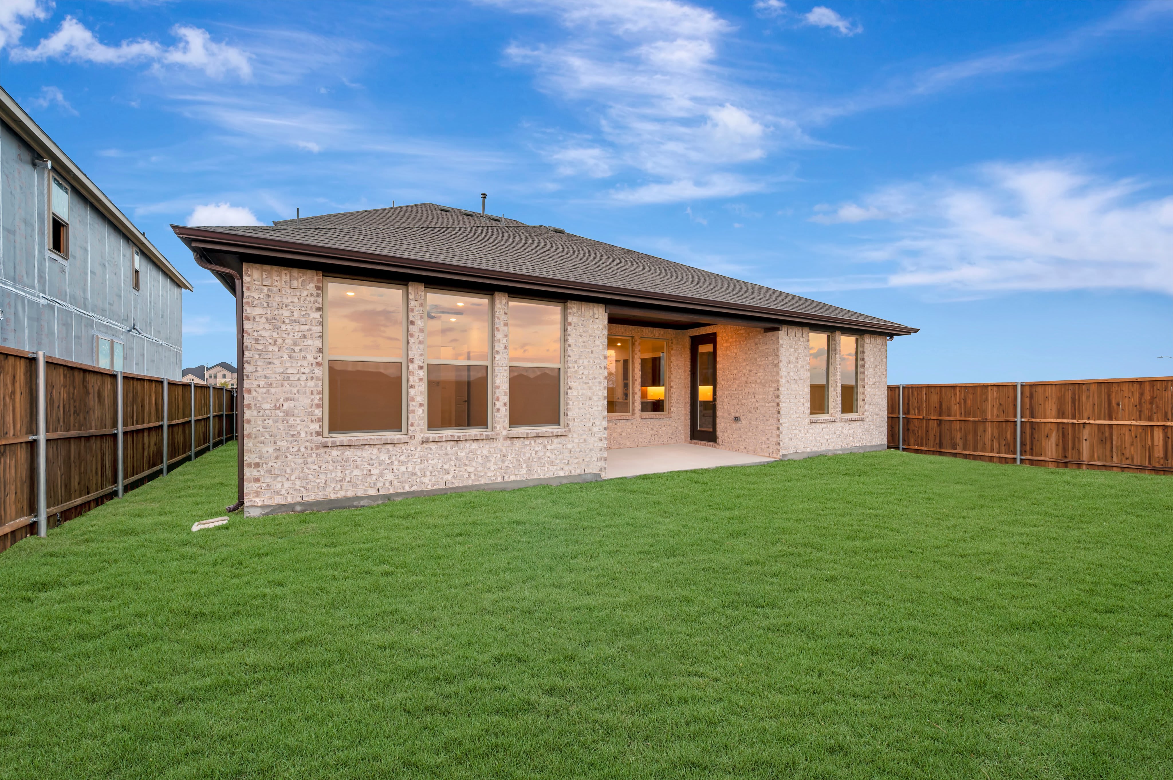 A lush green lawn fills the foreground, leading to a modern brick house set against a bright blue sky with wispy clouds.