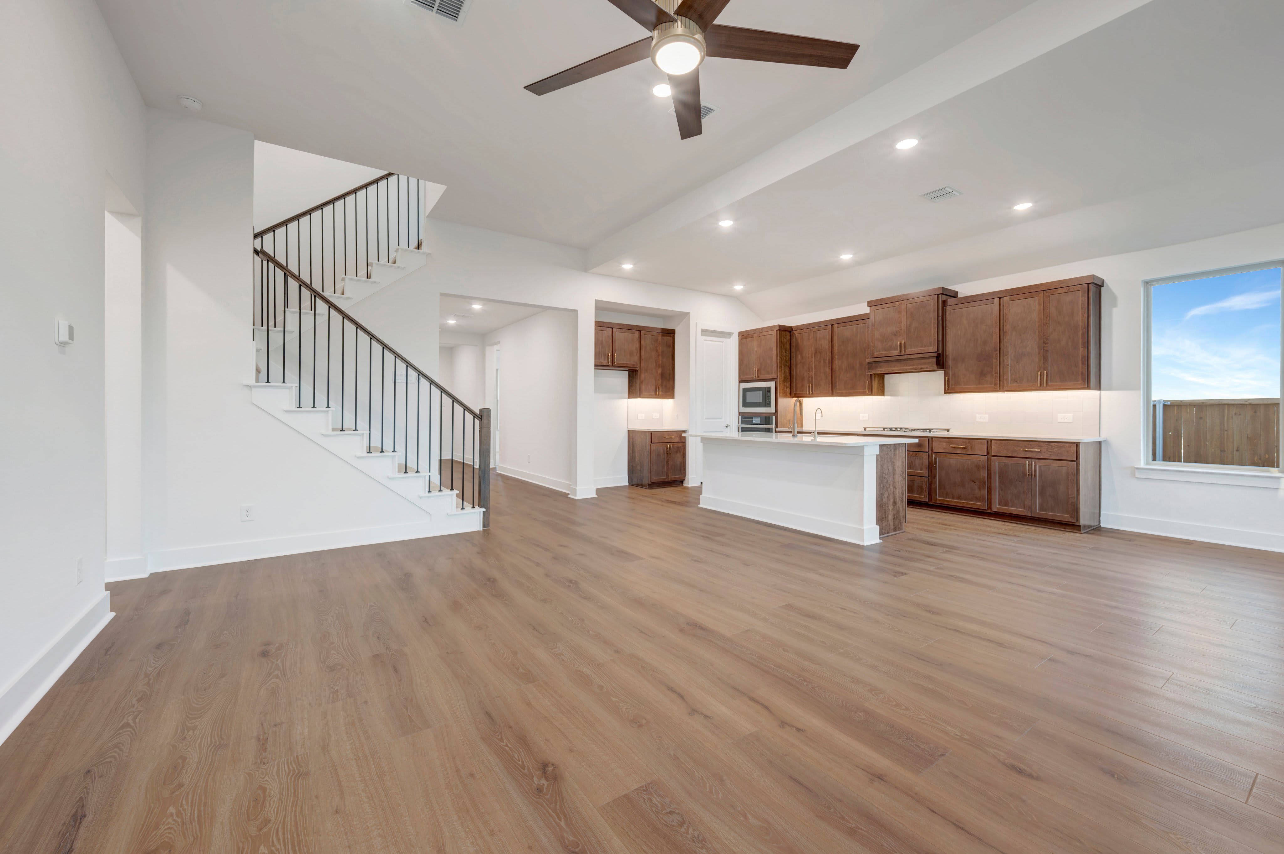 An open-concept living area with light wood flooring in the foreground leads to a kitchen with dark wood cabinets and a staircase in the background.