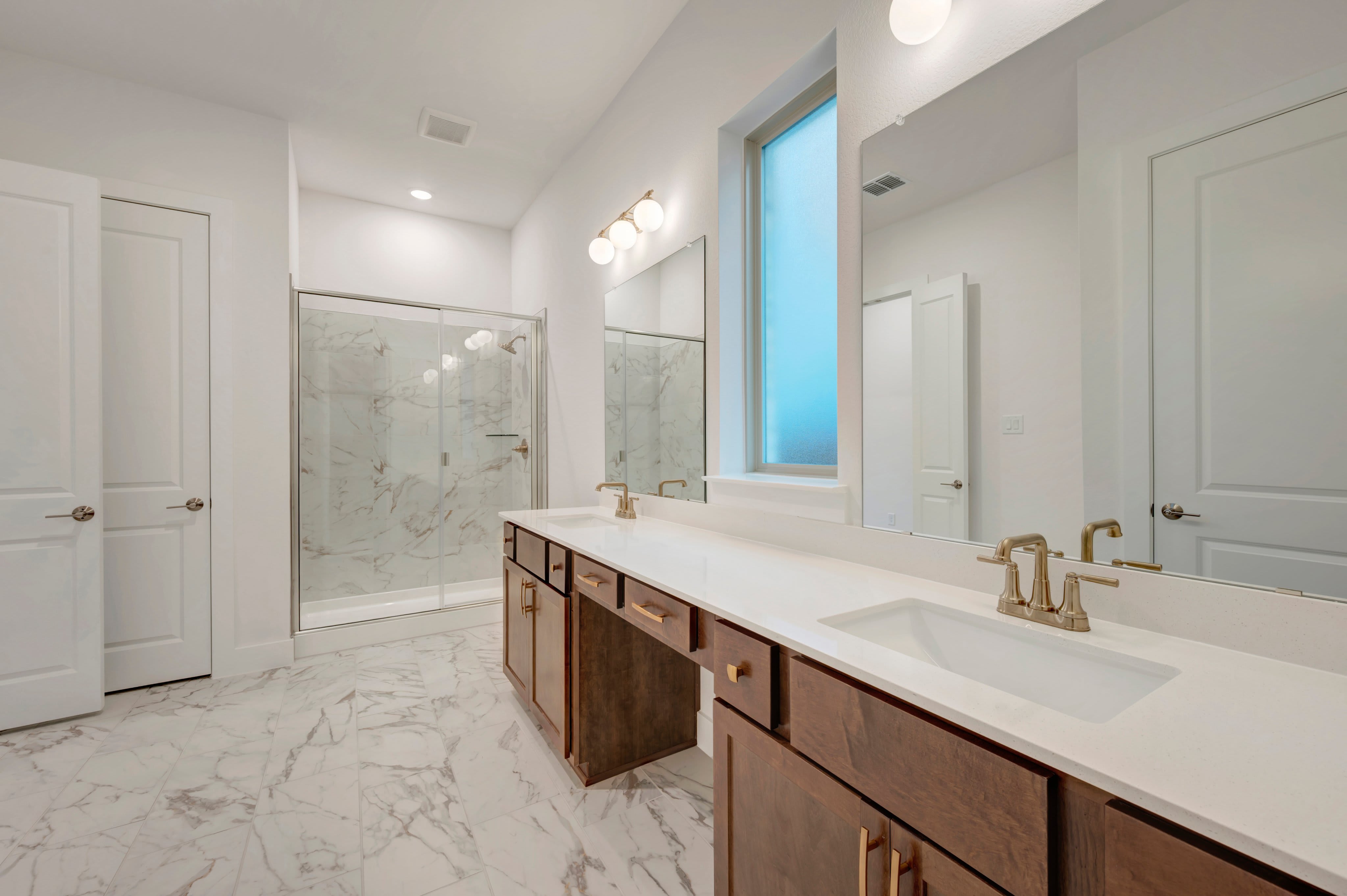 A modern bathroom features a double vanity with dark wood cabinets and white countertops in the foreground, with a glass-enclosed marble shower and white doors in the background.