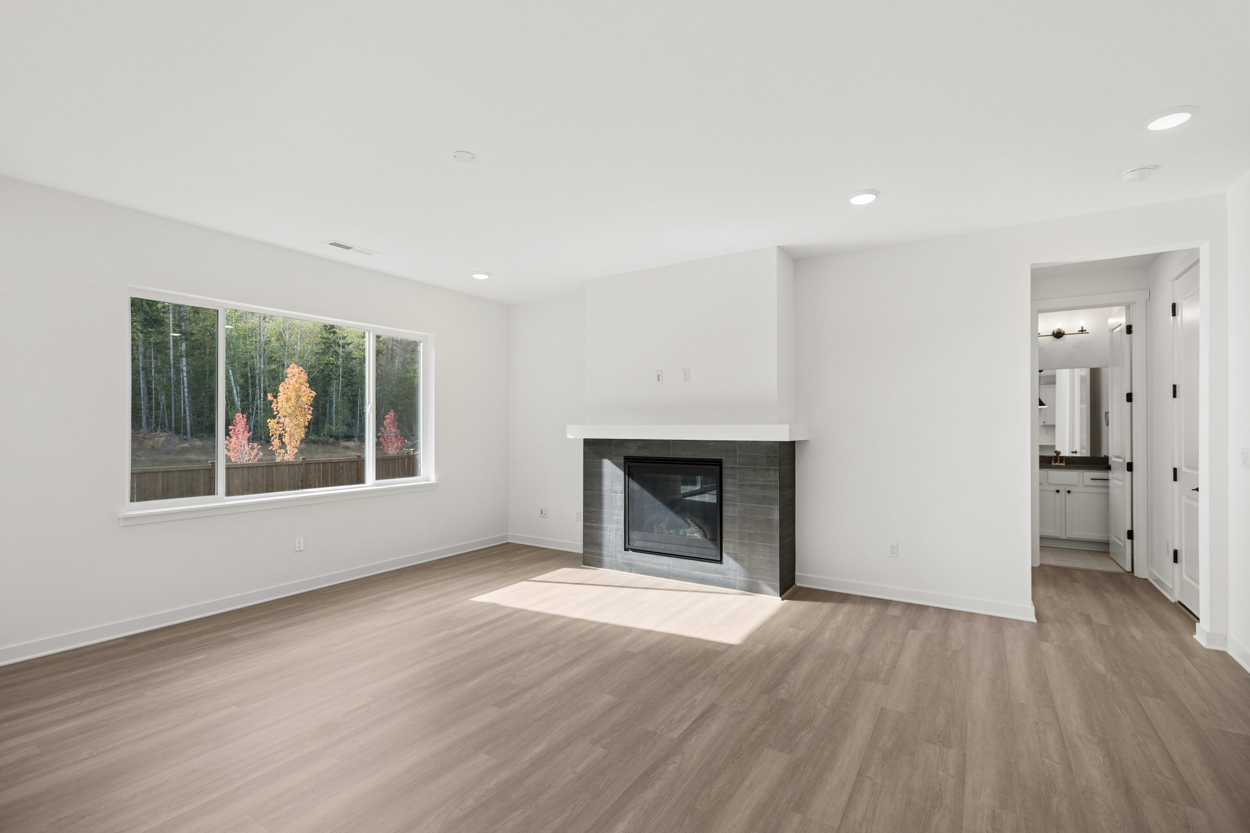 An empty room with light wood flooring and a modern fireplace in the foreground, with a large window overlooking a forest and a doorway leading to a bathroom in the background.