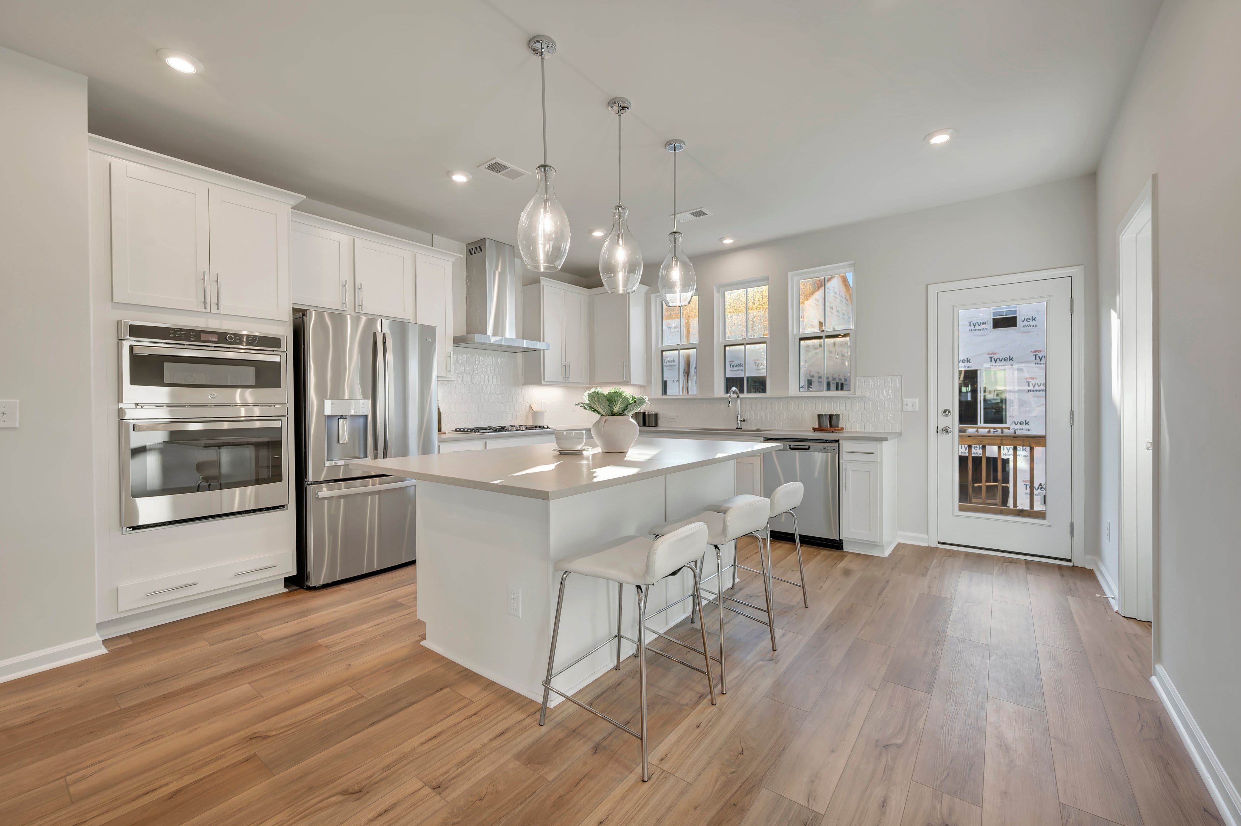 A modern white kitchen with a central island and barstools sits atop light wood flooring, featuring stainless steel appliances and a glass door in the background.