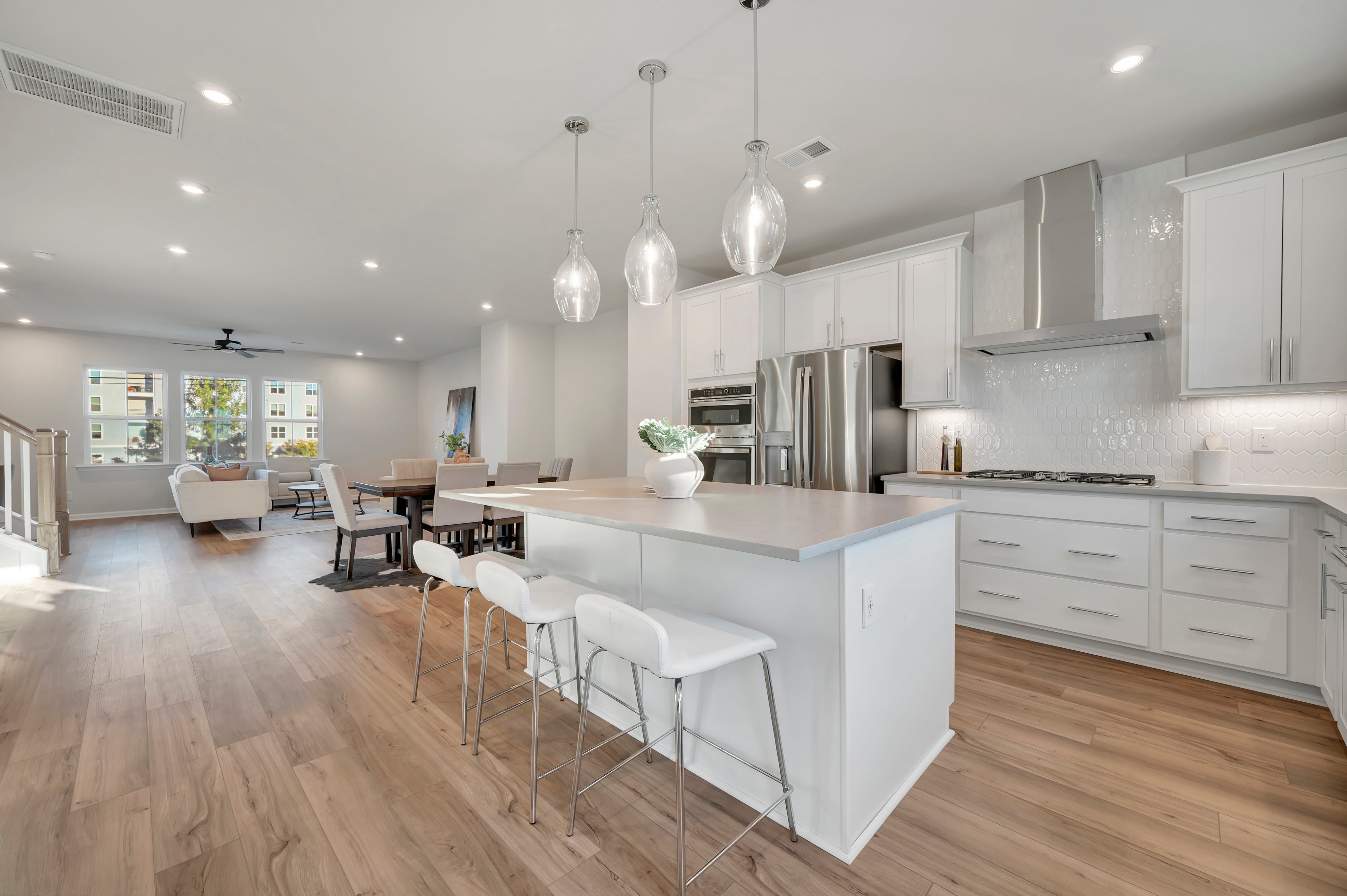 A modern white kitchen with a large island and barstools sits in the foreground, opening into a bright living and dining area in the background.