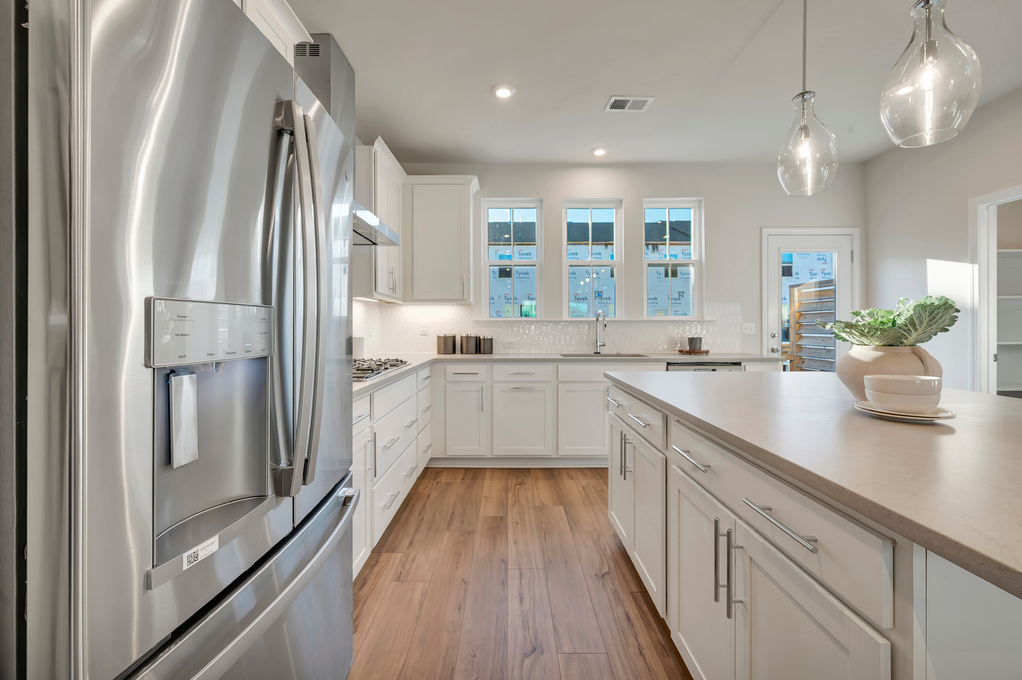 A stainless steel refrigerator stands in the foreground of a modern white kitchen with a large center island and hardwood floors.