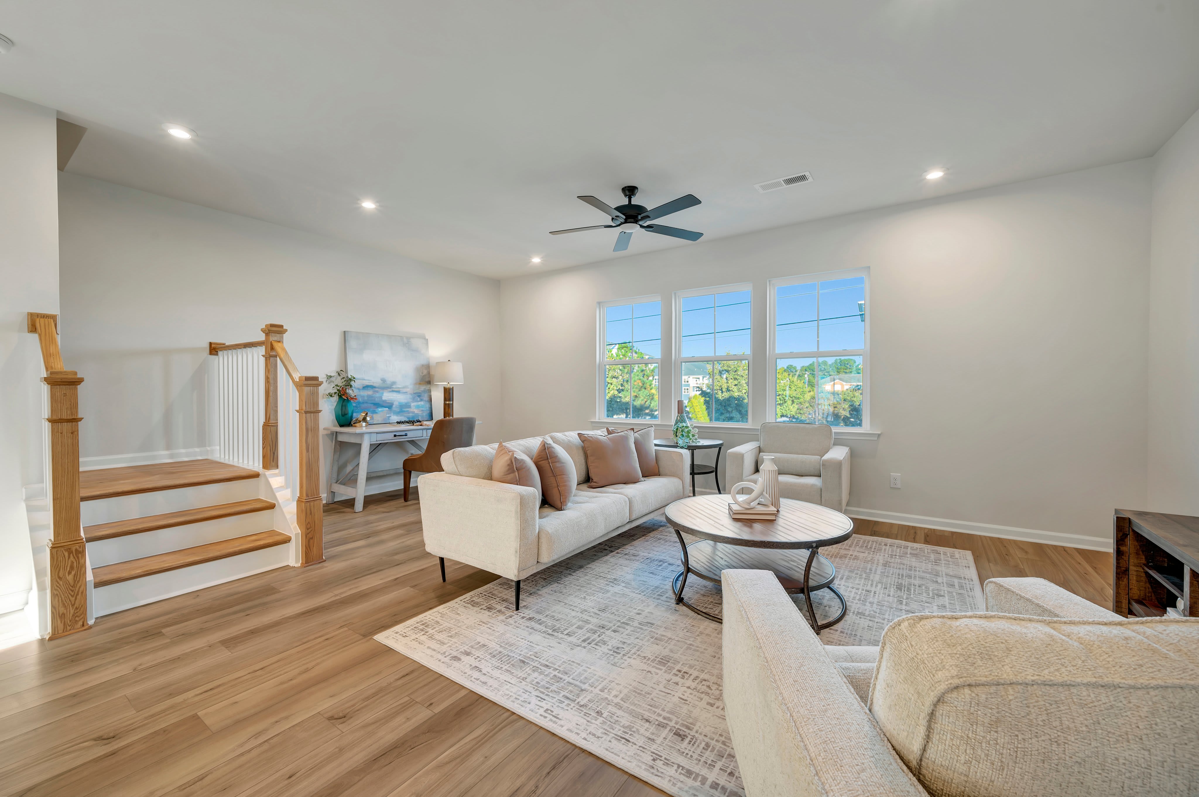 A modern living room features a light-colored sofa and coffee table on a patterned rug in the foreground, with a wooden staircase to the left and large windows overlooking greenery in the background.