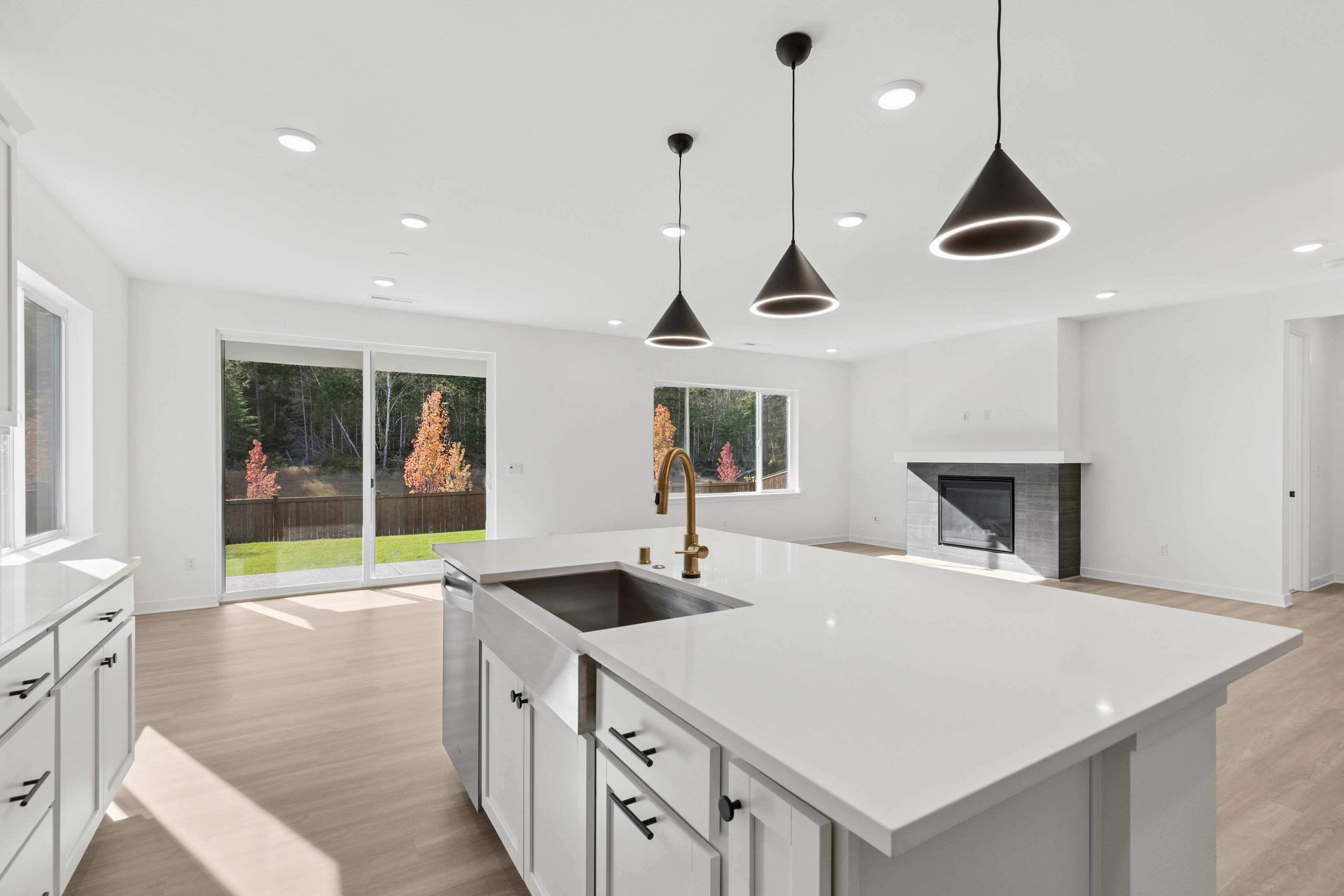 A modern white kitchen island with a gold faucet sits in the foreground of a bright, open-concept living space featuring a fireplace and sliding glass doors leading to a backyard.