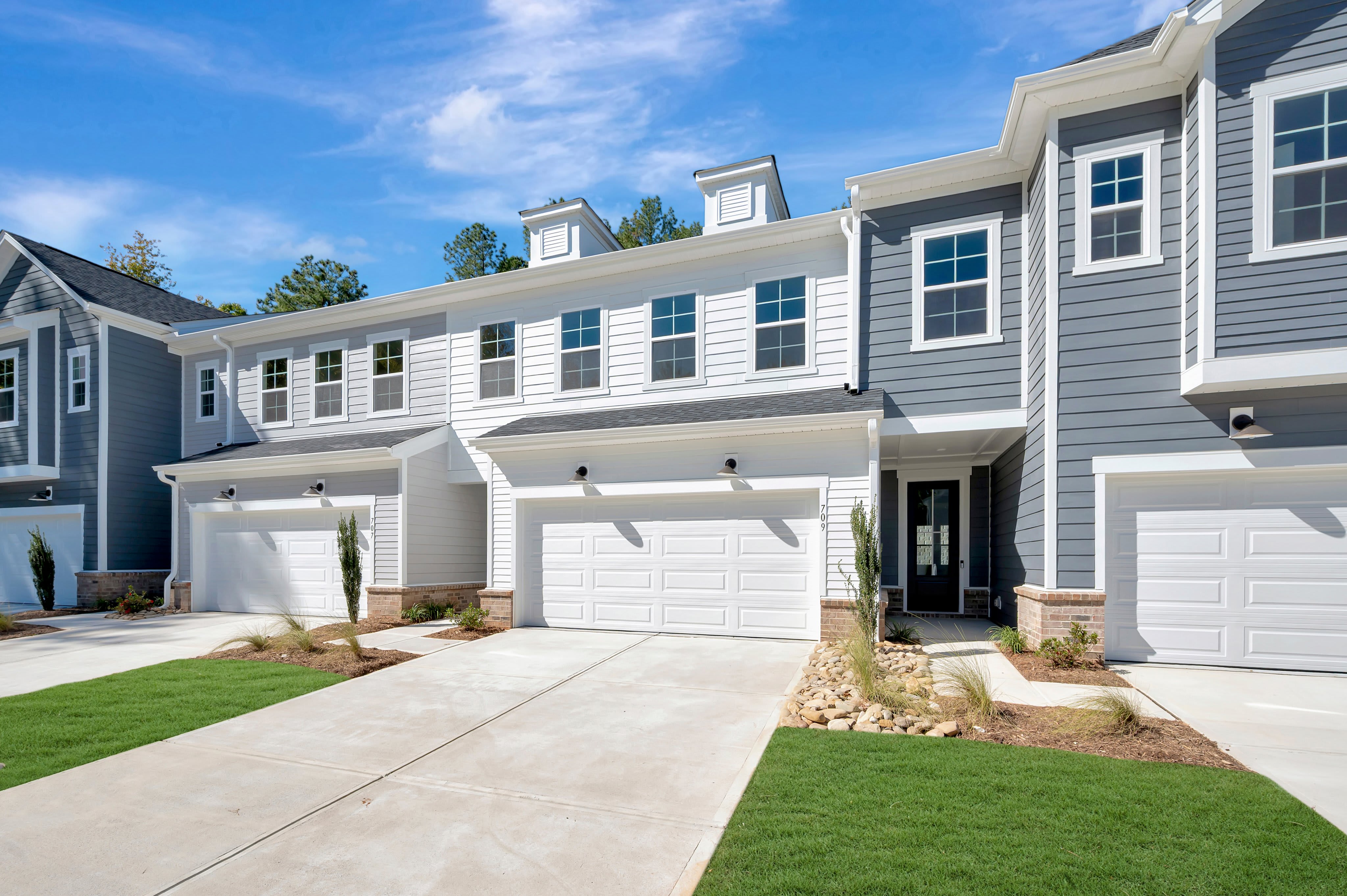 Concrete driveways and green lawns lead up to a row of modern grey and white townhomes under a bright blue sky.