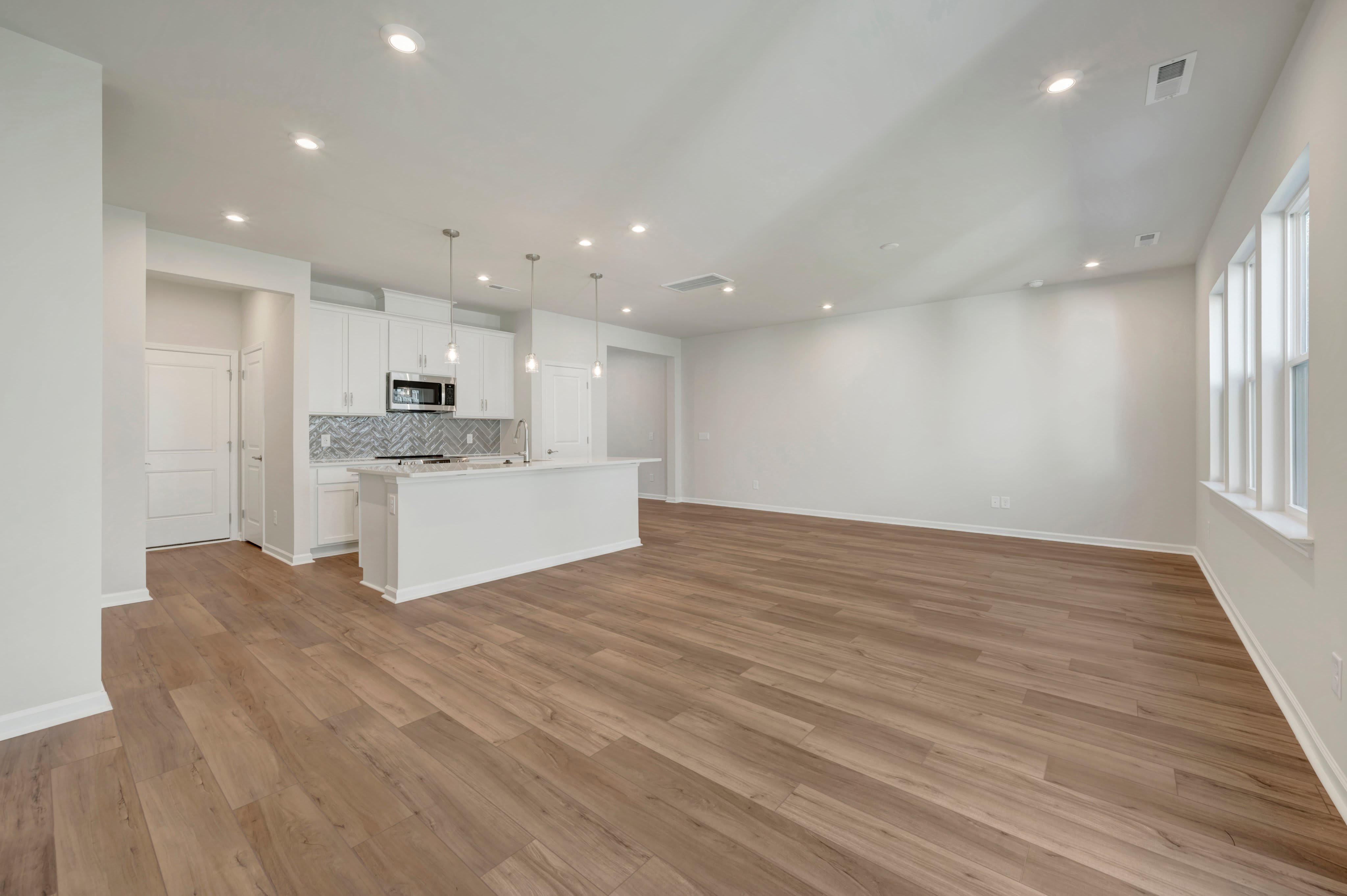 A wide-planked wooden floor fills the foreground, leading back to a modern white kitchen and an open living area with white walls.