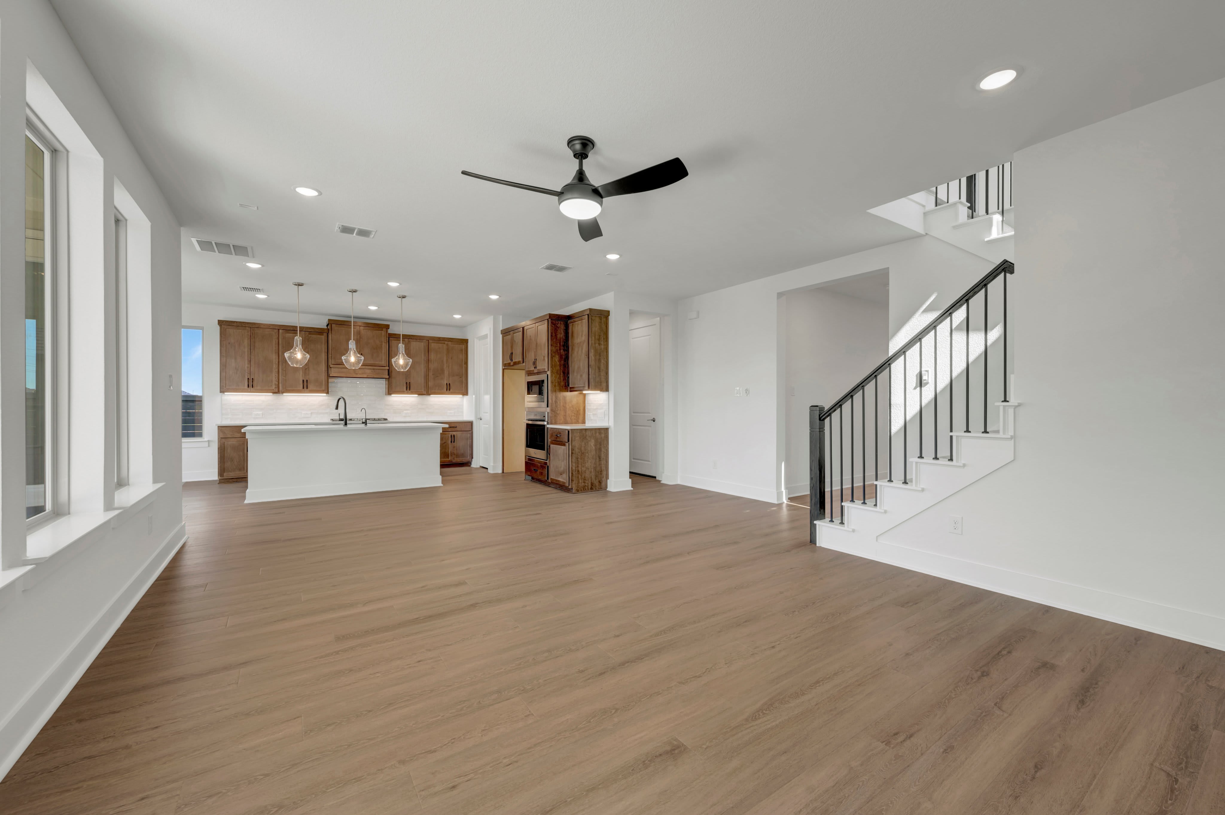 An open-concept living area with light wood flooring in the foreground leads to a modern kitchen and a white staircase in the background.