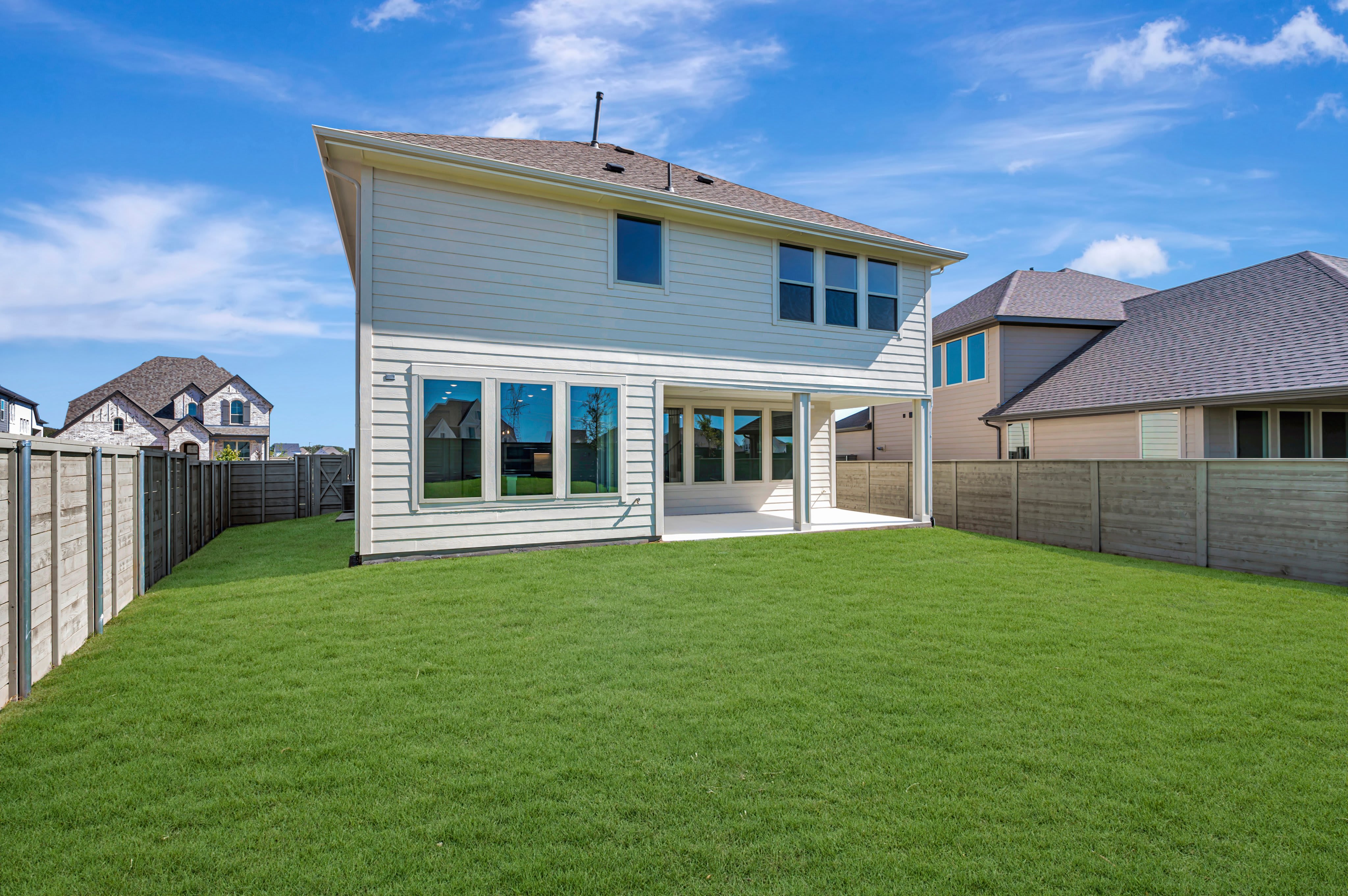 A lush green lawn fills the foreground, leading to a two-story white house set against a bright blue sky with wispy clouds and neighboring homes.