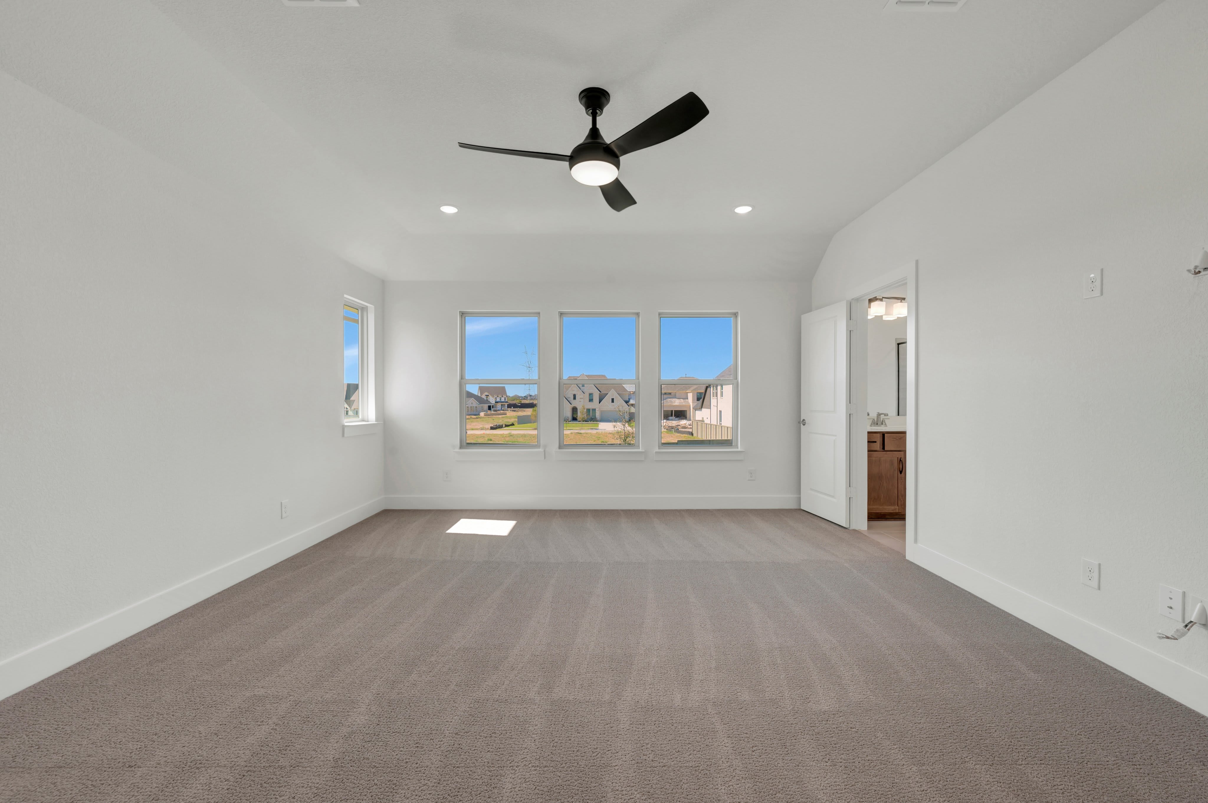 An empty room with beige carpeting and white walls features a black ceiling fan in the foreground and a view of suburban houses through three windows in the background.