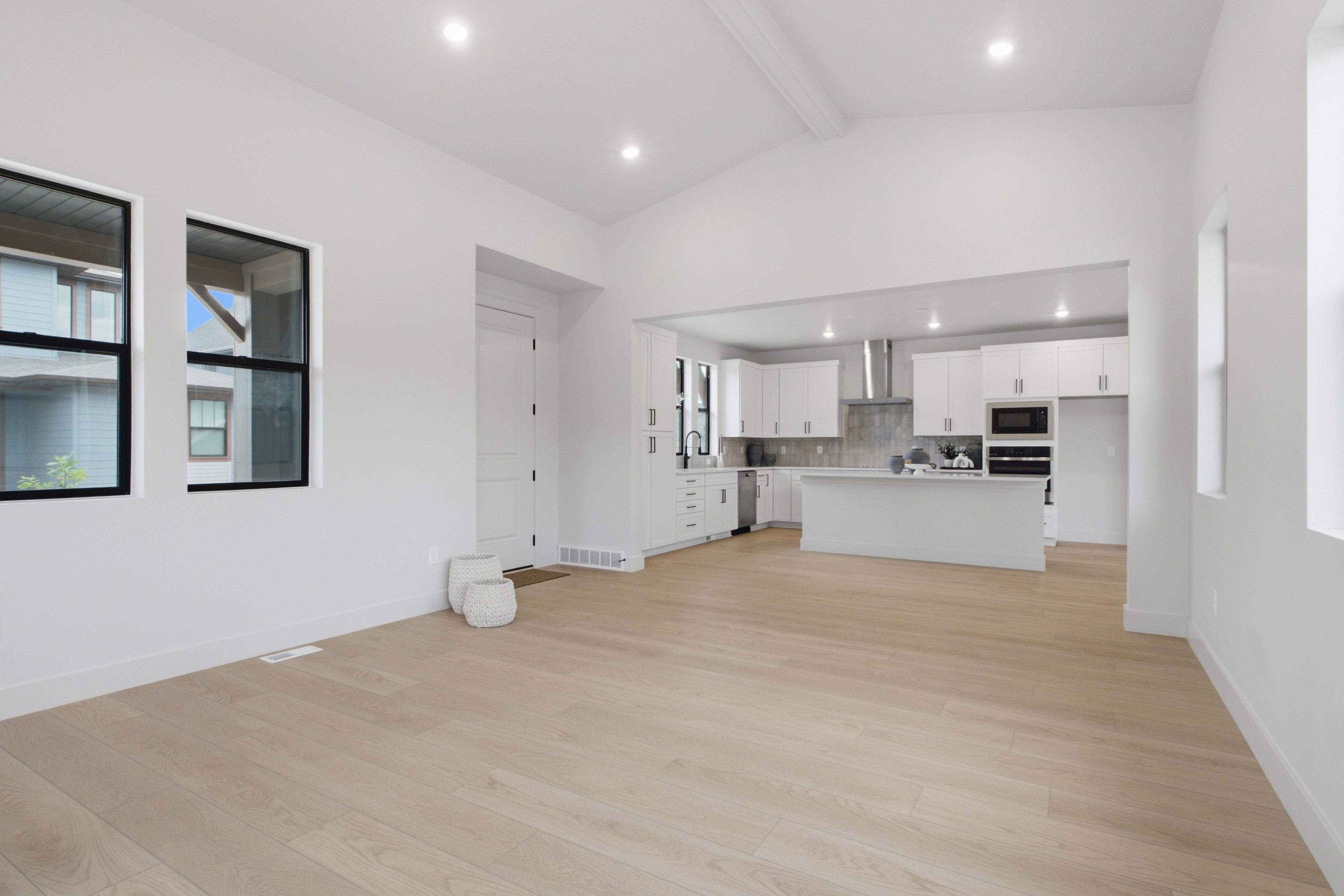 An empty room with light wood flooring leads into a modern white kitchen in the background.
