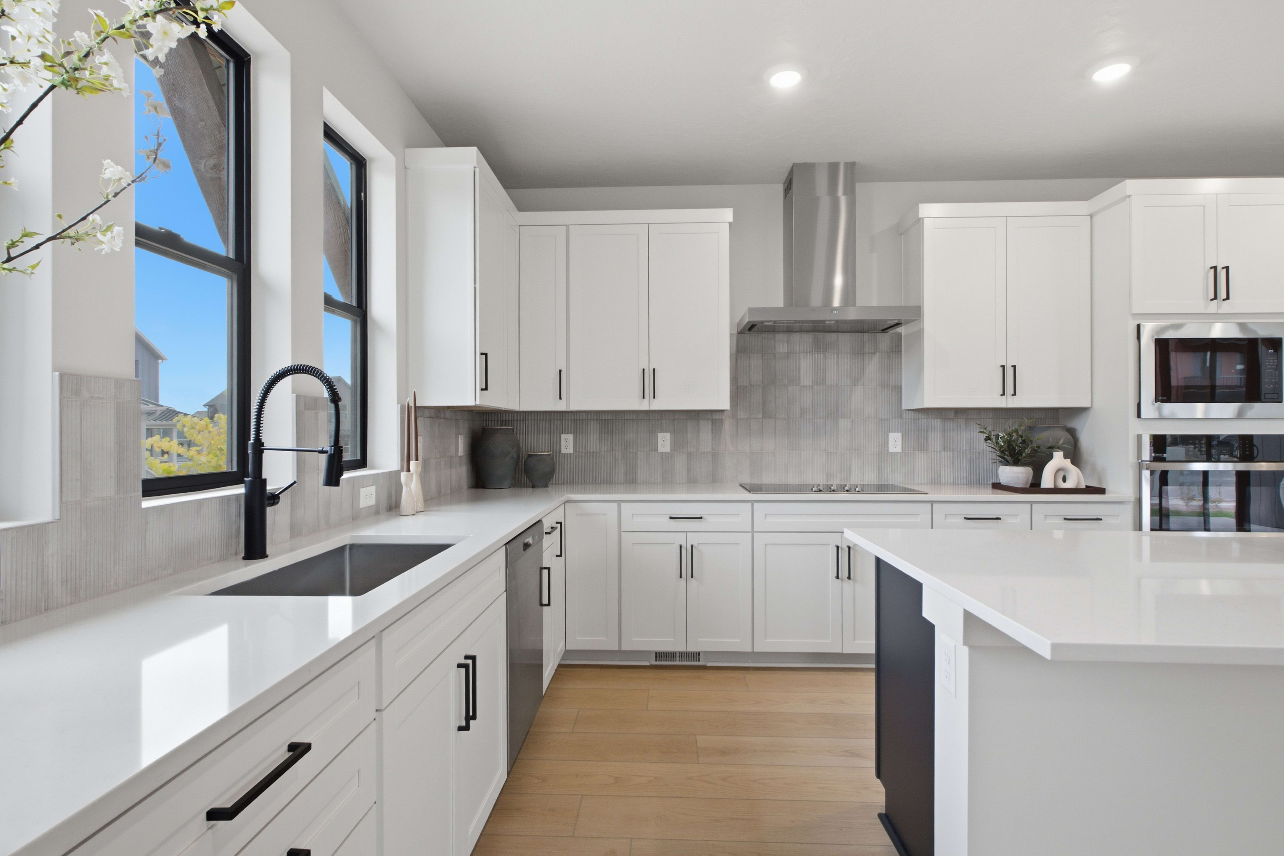A modern white kitchen features a sleek island and sink in the foreground, with white cabinetry, stainless steel appliances, and a tiled backsplash in the background.