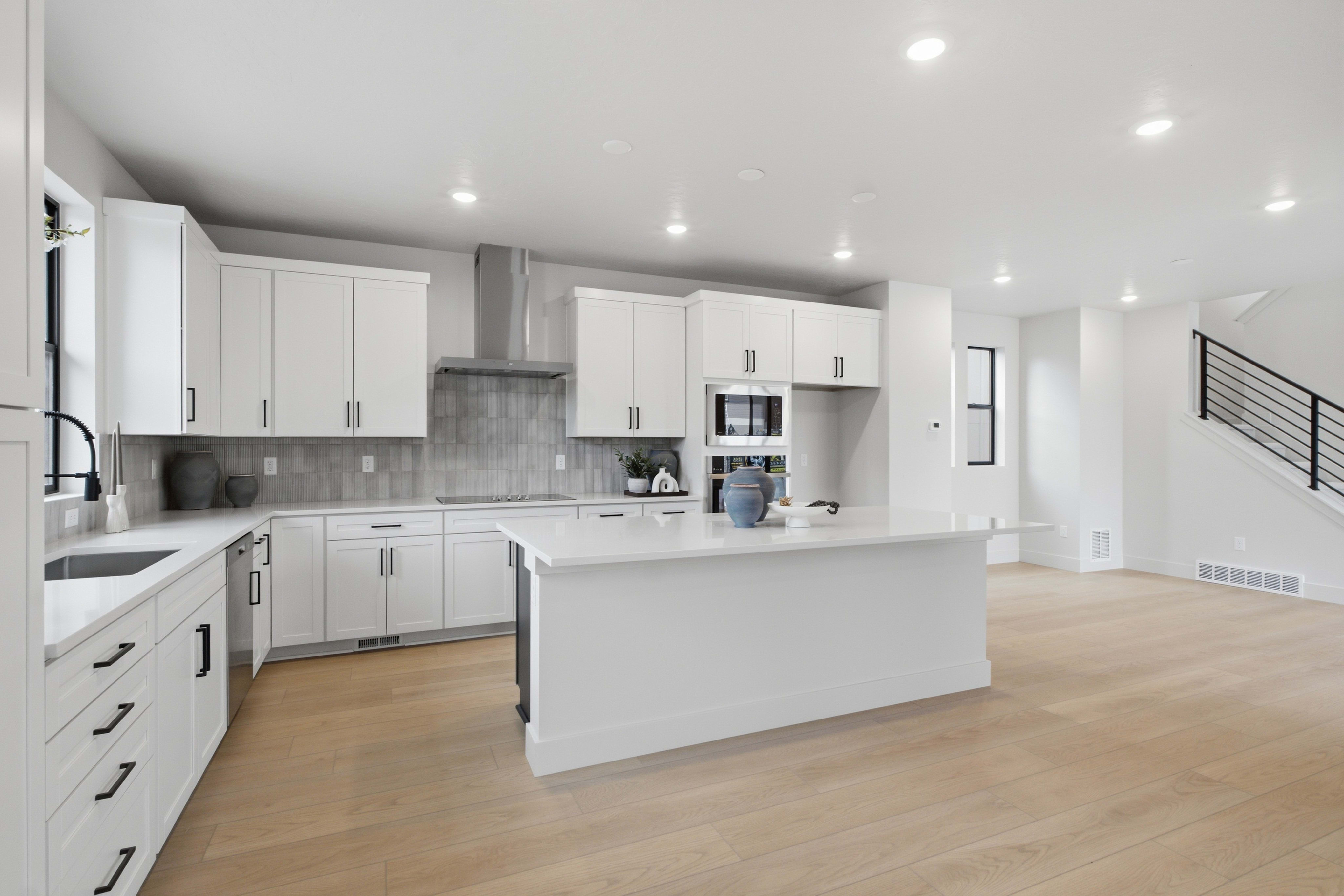 A modern white kitchen with a large center island and light wood flooring sits in the foreground, while a white hallway and staircase are visible in the background.