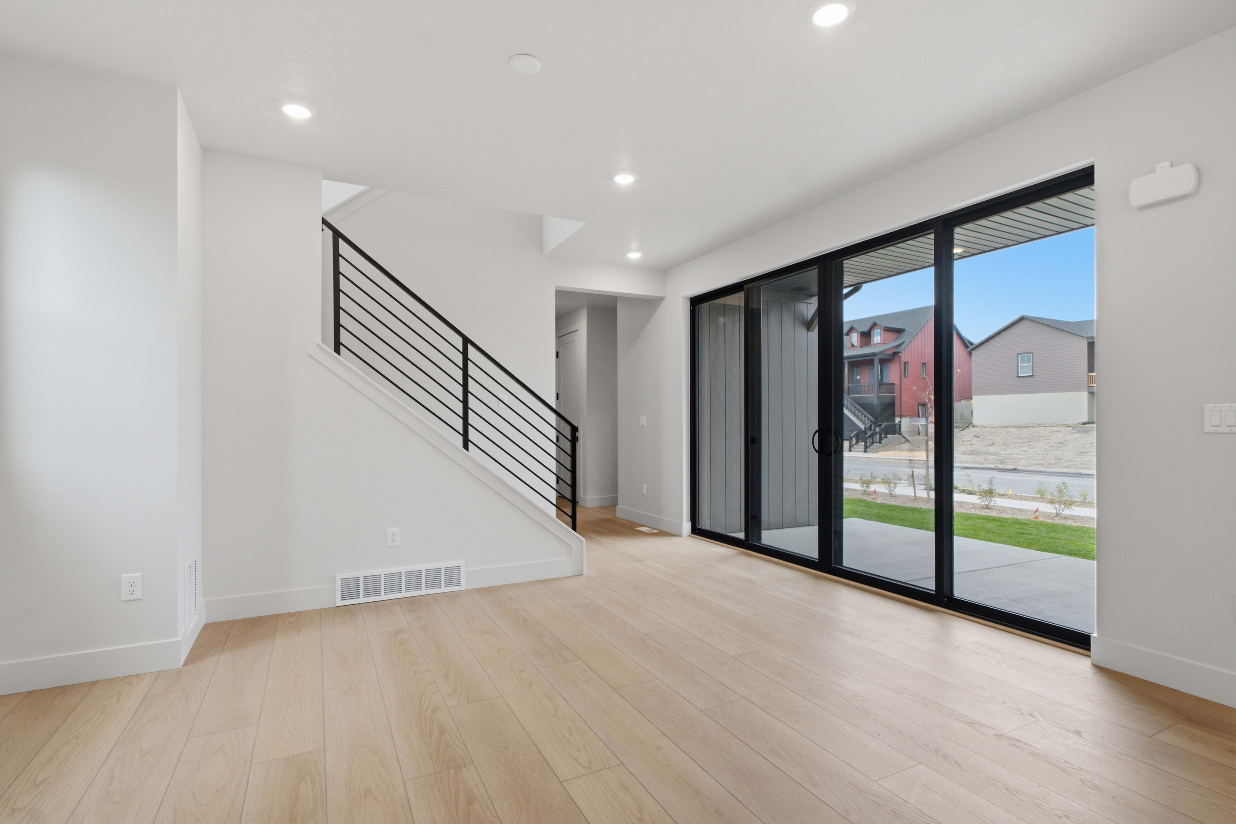An empty room with light wood floors and a modern staircase in the foreground opens up to a view of neighboring houses through large sliding glass doors in the background.