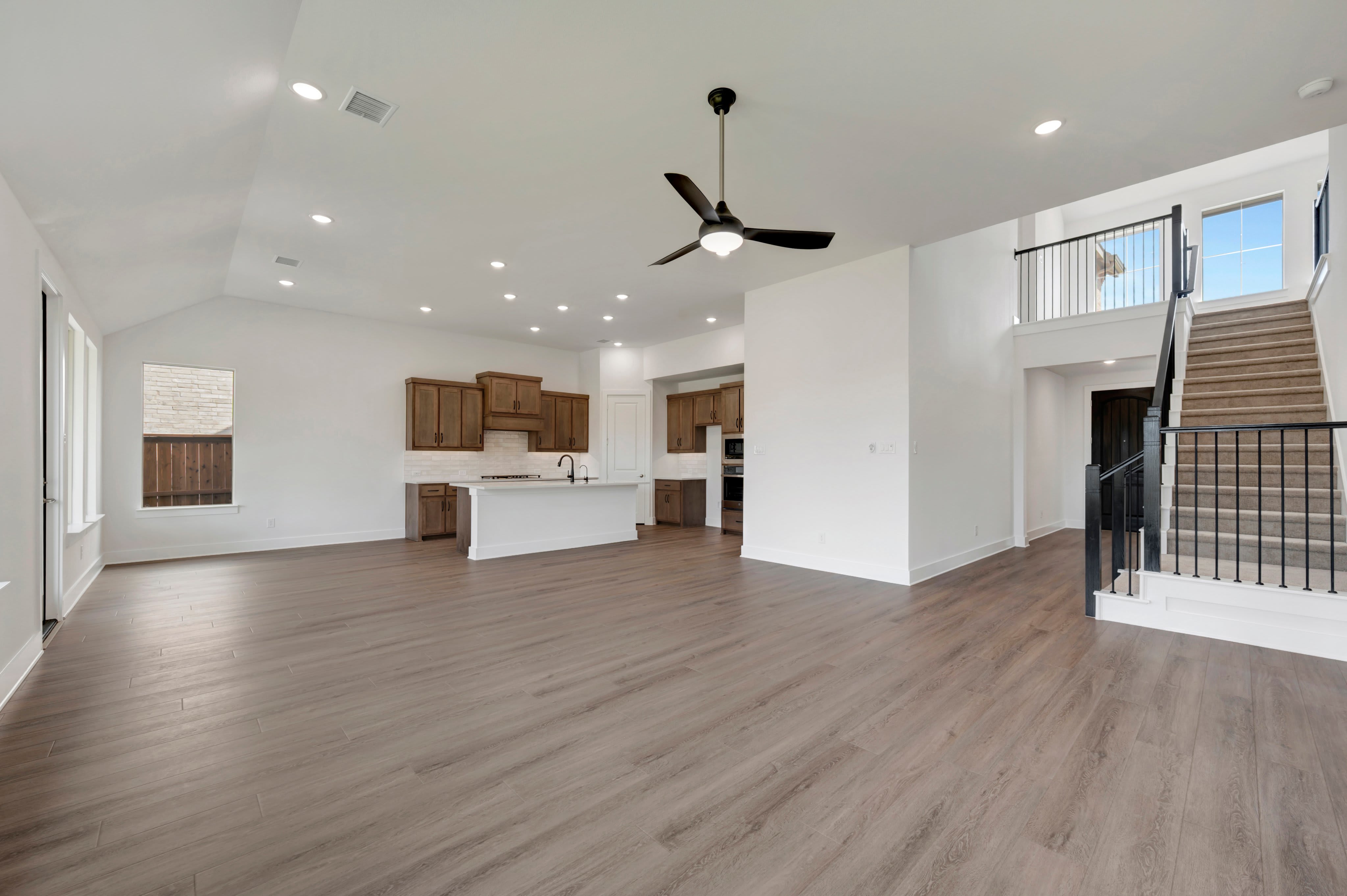 An open-concept living area with grey wood flooring in the foreground leads to a kitchen with wooden cabinets and a staircase in the background.