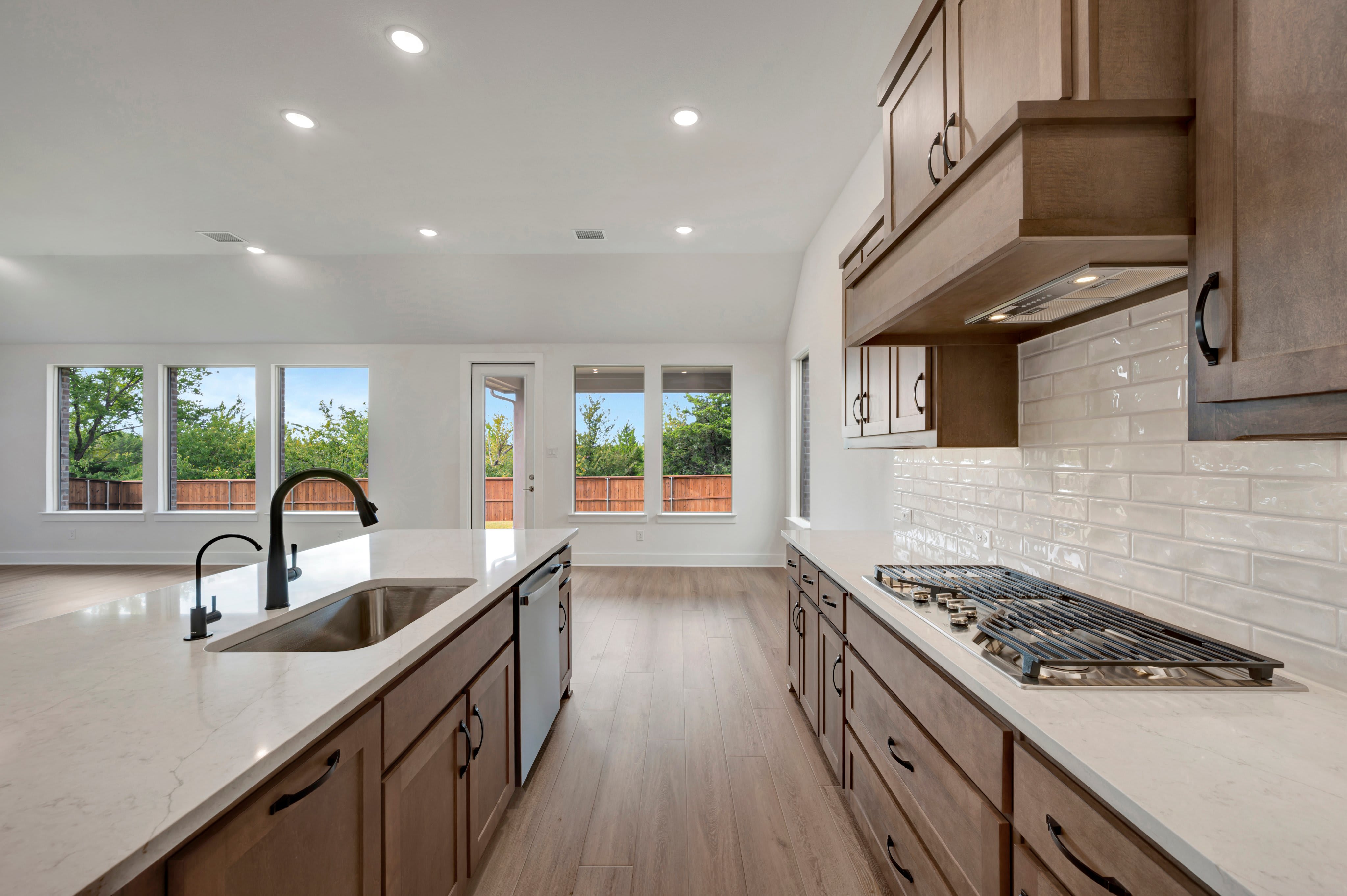 A modern kitchen features a white quartz island with a black faucet in the foreground and a gas stovetop with wooden cabinetry and large windows in the background.