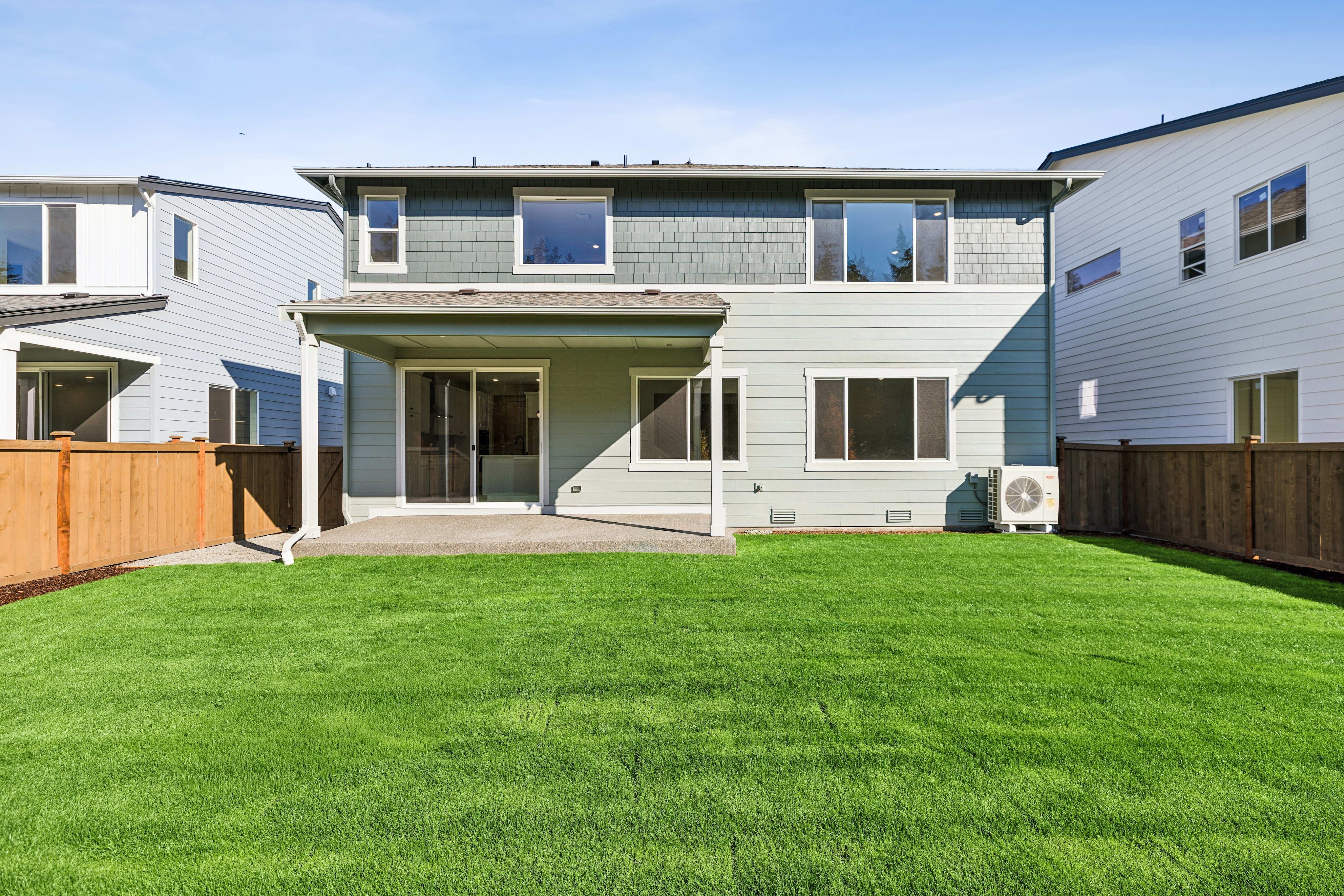 A lush green lawn fills the foreground, leading to a light-colored two-story house with a covered patio and neighboring homes in the background.