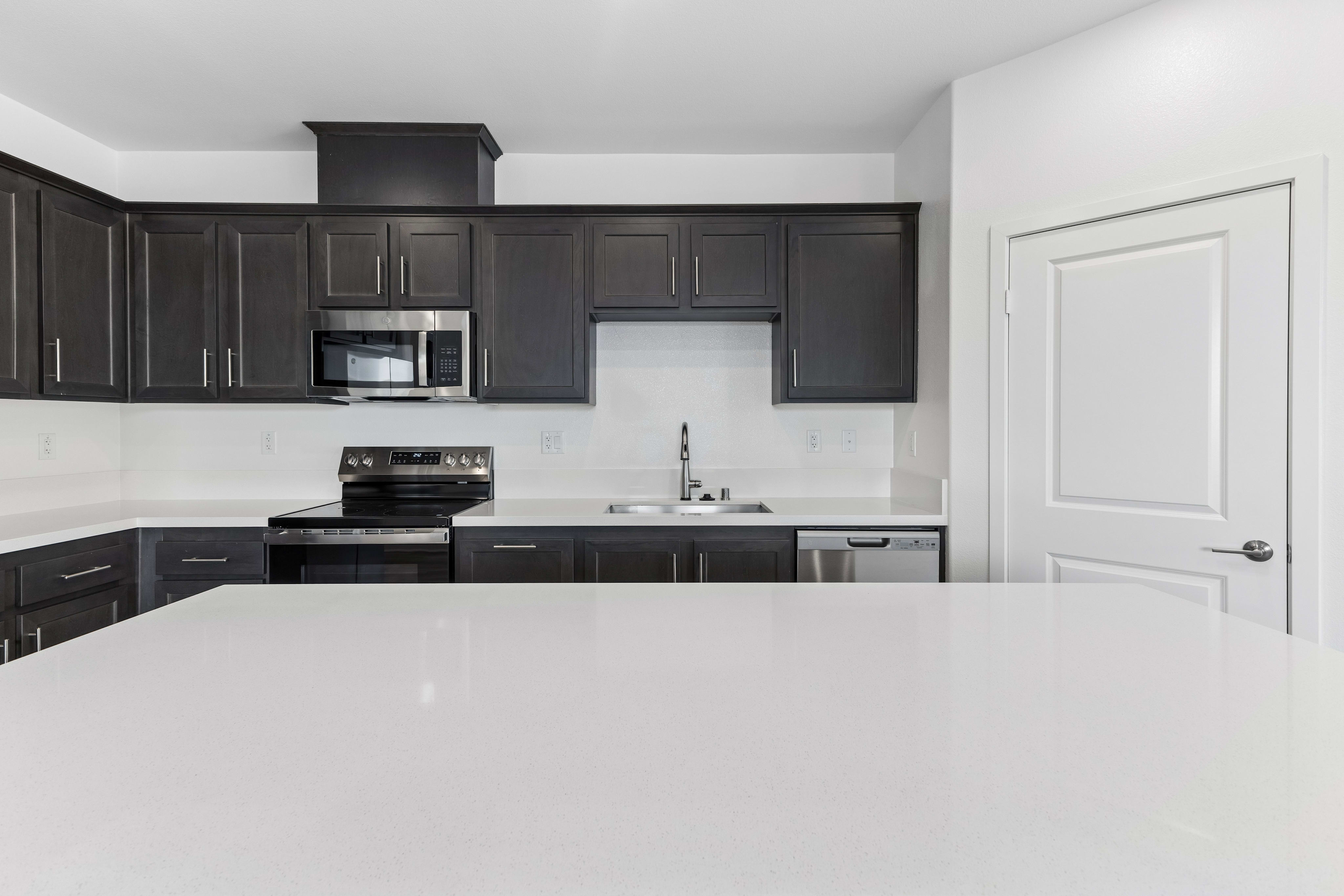 A white countertop sits in the foreground of a modern kitchen featuring dark brown cabinetry, stainless steel appliances, and a white door in the background.