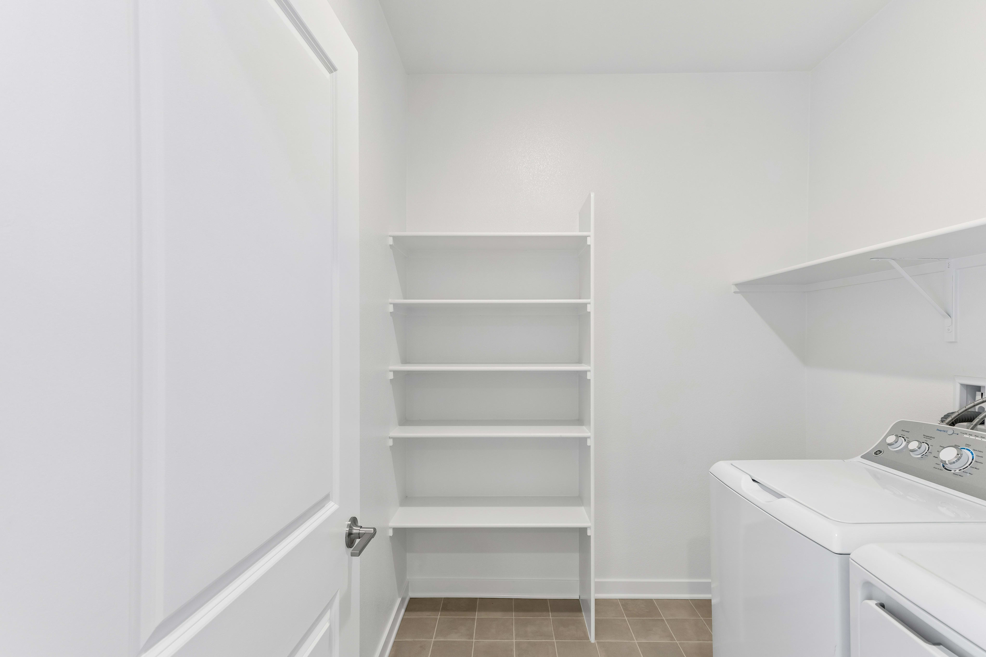 A white door stands in the foreground of a laundry room featuring white shelving and a washing machine in the background.