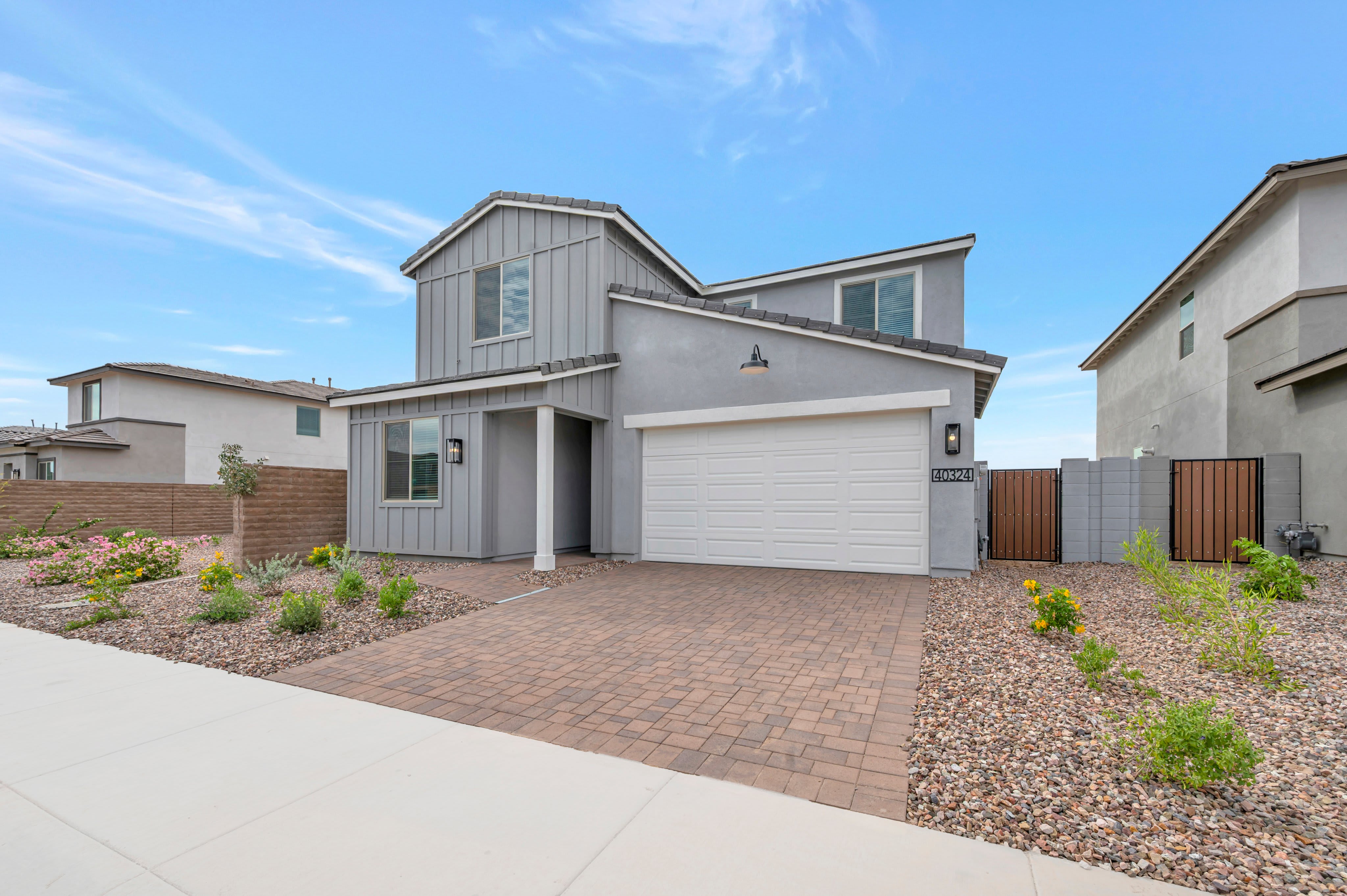 A modern grey house with a white garage door and brick driveway sits under a blue sky, surrounded by desert landscaping and neighboring homes.