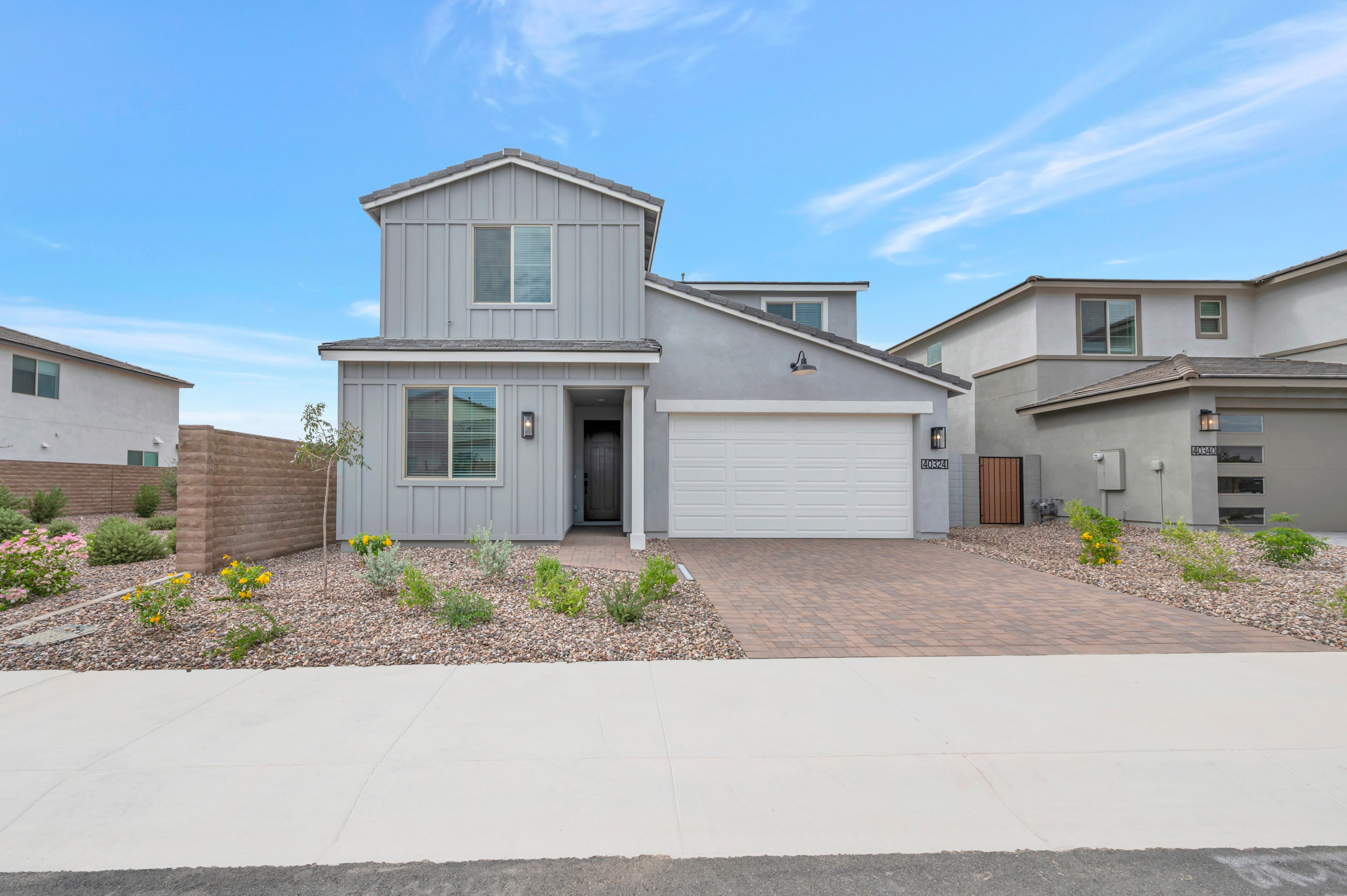 A modern grey two-story house with a white garage door sits behind a landscaped gravel yard and a concrete sidewalk under a bright blue sky.