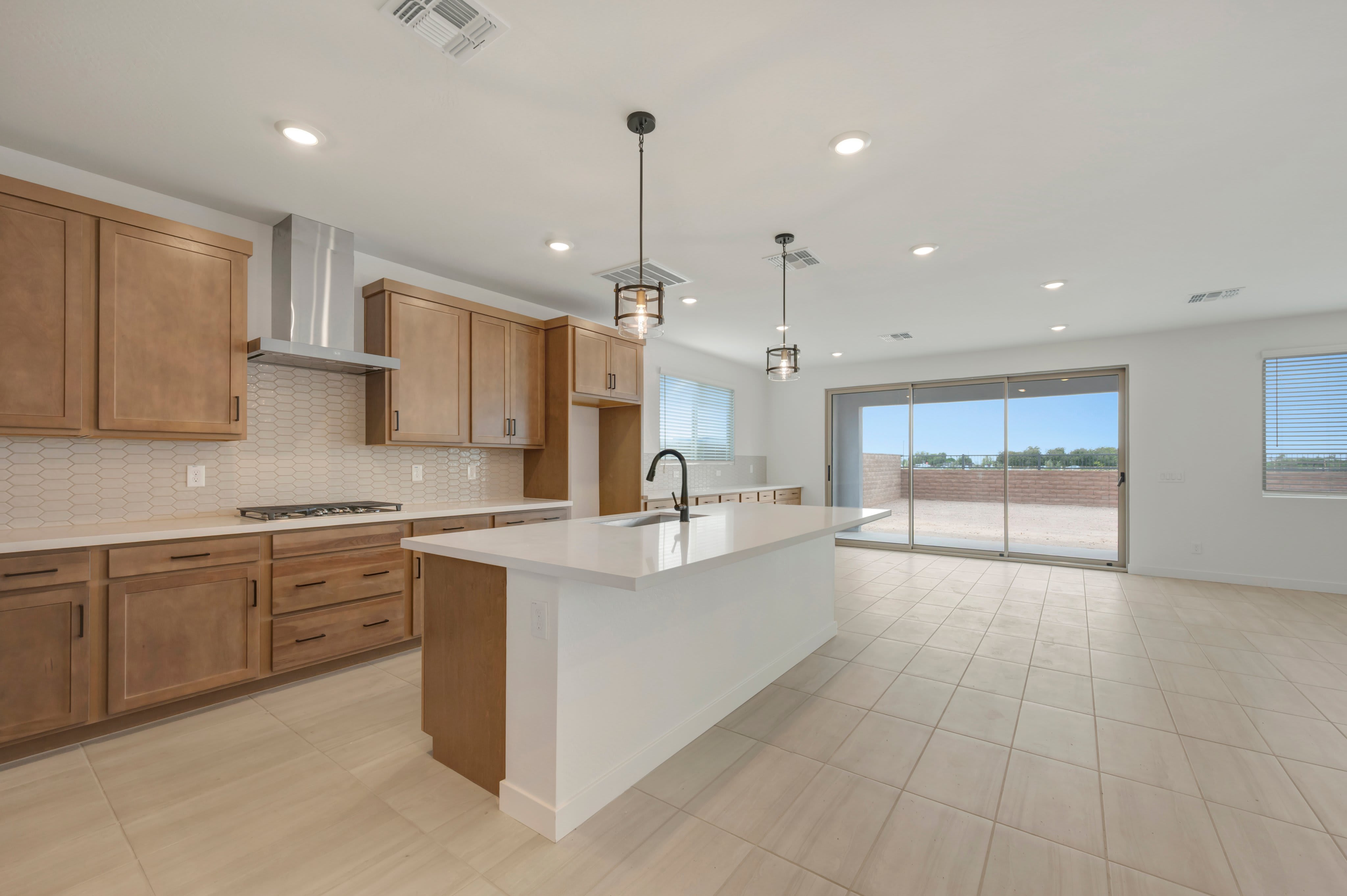 A modern kitchen with light wood cabinets and a white island sits in the foreground, leading to a bright living area with sliding glass doors in the background.